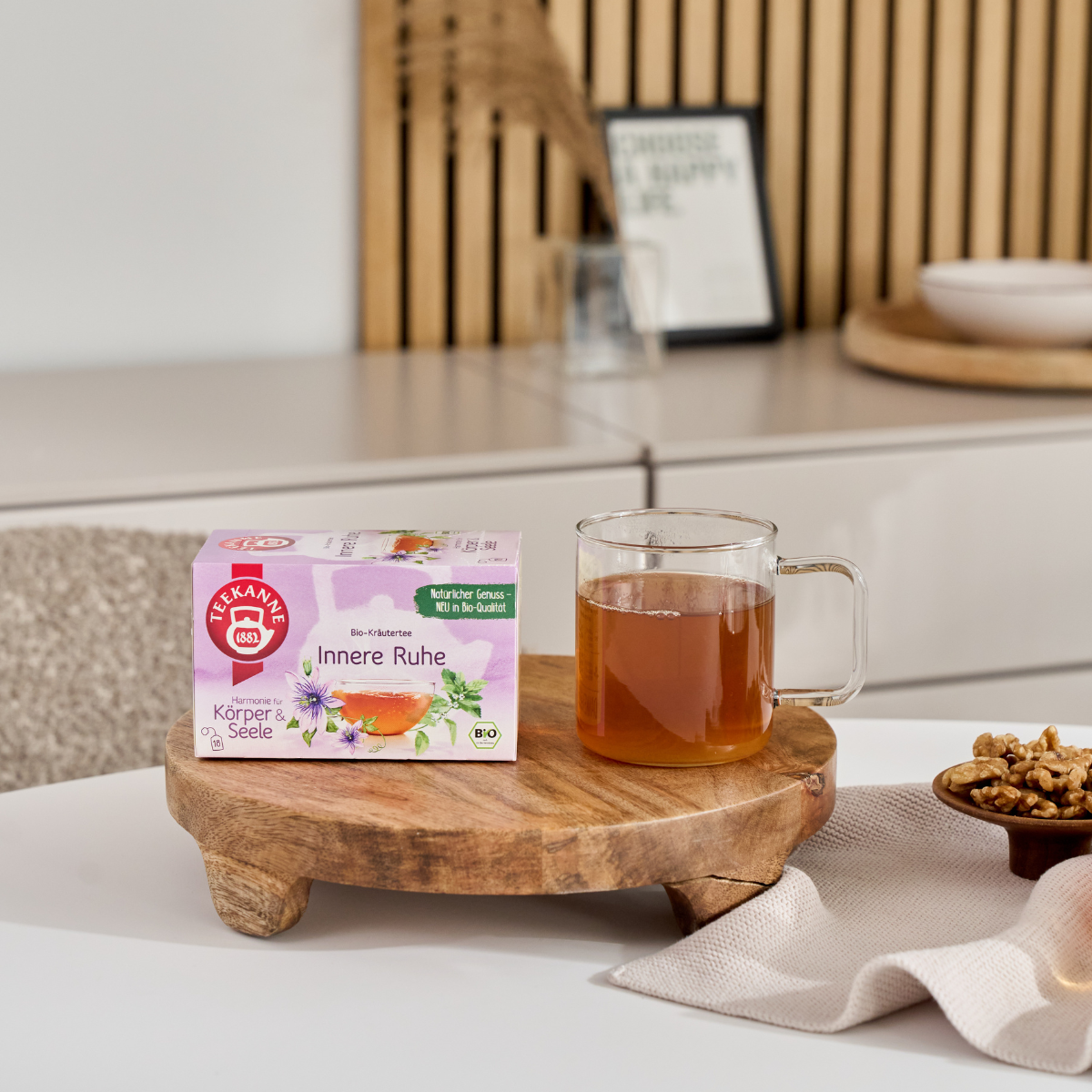Ein Glasbecher „BIO Innere Ruhe“ und die dazugehörige Schachtel stehen auf einem Holzständer, während in der Nähe eine kleine Schale mit Walnüssen auf einem beigen Tuch in einer modernen, minimalistischen Küchenumgebung steht.