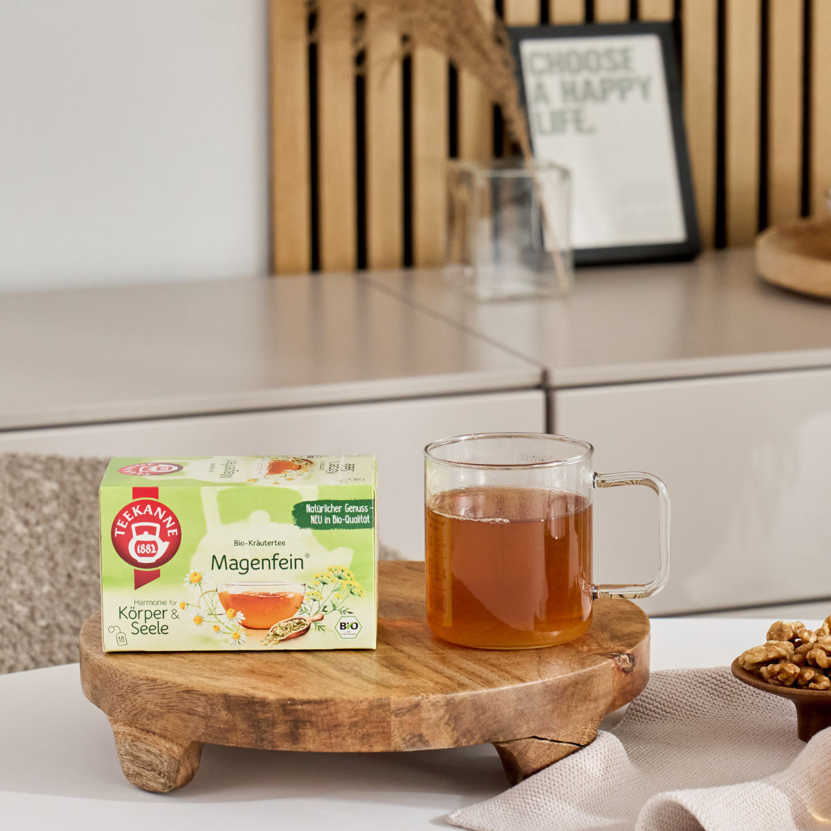 Eine Glastasse Tee und eine Schachtel BIO Magenfein® stehen auf einem Holztablett mit Walnüssen und Wohndekor, umrahmt von einem gemütlichen, modernen Ambiente, das diese wohltuende Kräuterteemischung mit Kamille hervorhebt.