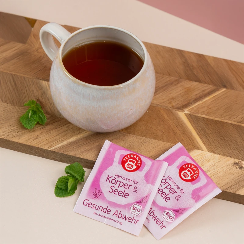 Auf einem Holzbrett steht eine Tasse Tee mit frischer Minze und zwei rosafarbenen Mix & Match-Päckchen „Harmonie für Körper & Seele“ – ideal für alle, die harmonisierende Aromen oder ein besonderes Angebot suchen.