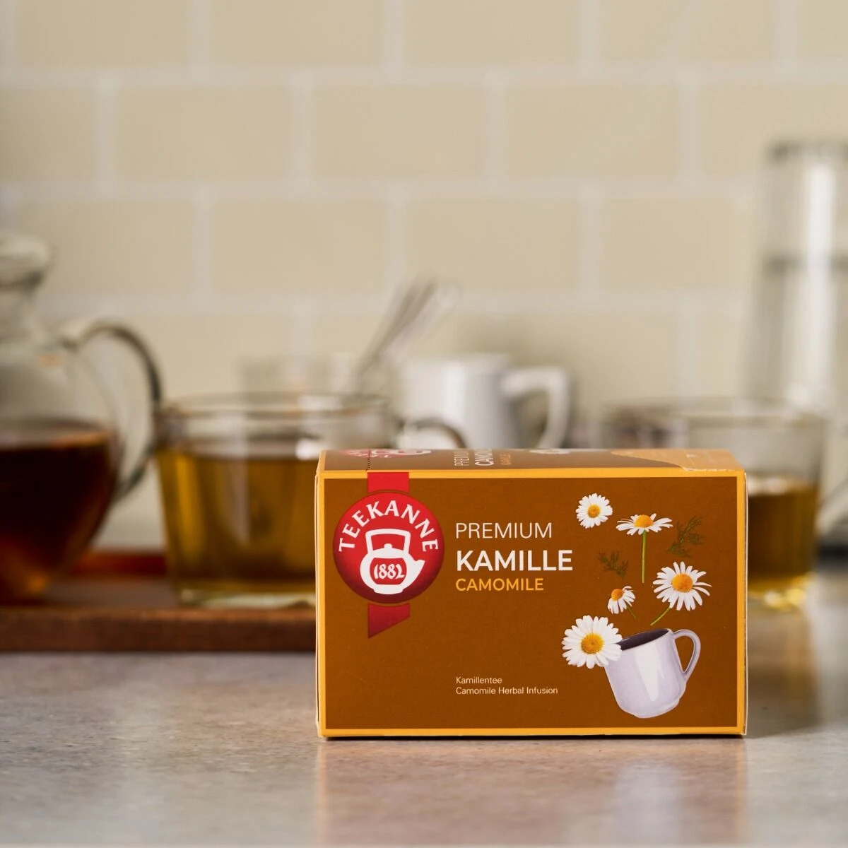 Eine Schachtel Premium Kamille Tee steht auf einem Küchentisch, begleitet von Kamillenblüten, einer Teekanne und Tassen. Die orangefarbene Verpackung zeigt beruhigende Blumen- und Teetassenmotive, die die beruhigende Essenz von Premium Kamille widerspiege