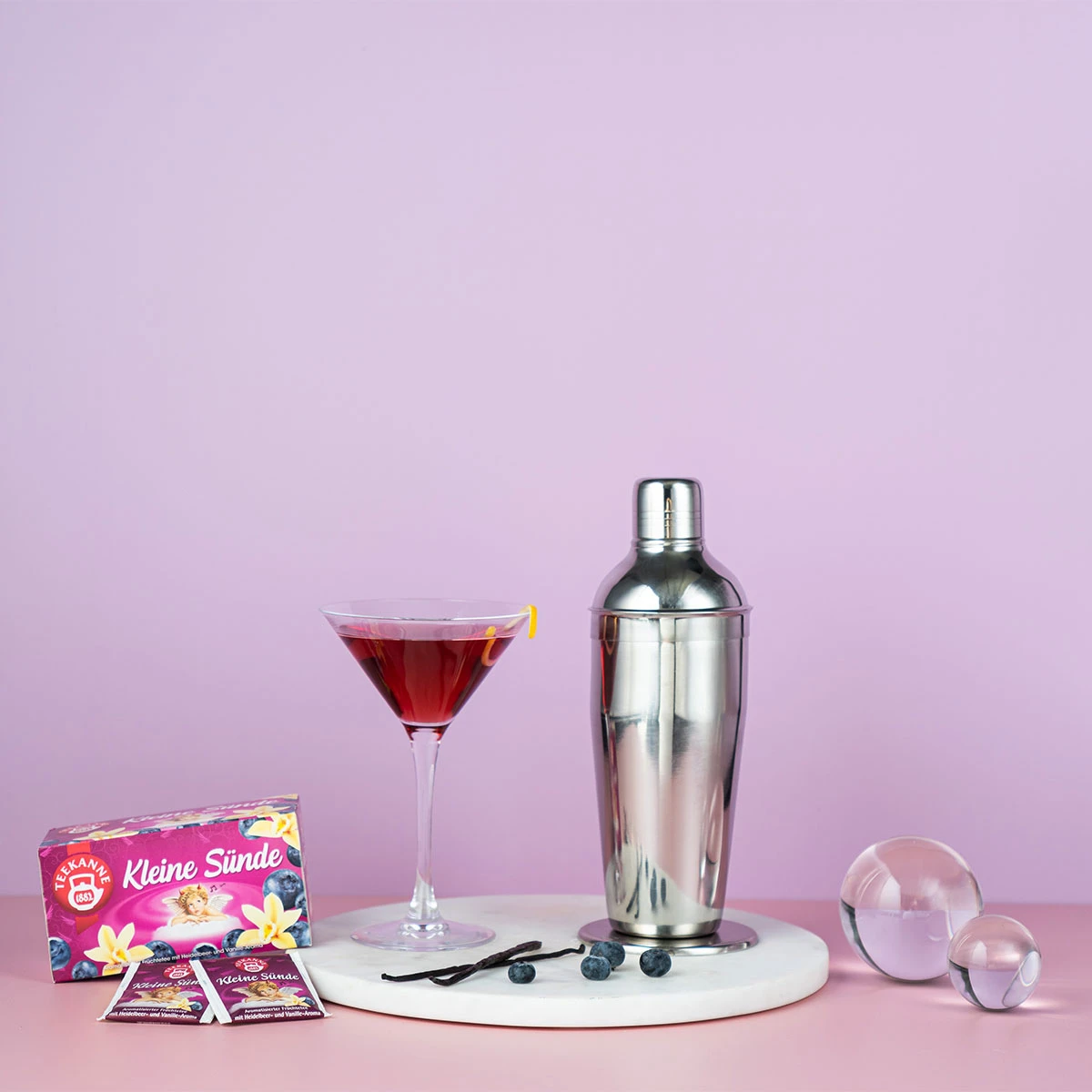 Ein rosafarbener Cocktail in einem Martiniglas mit Zitronendrehung steht neben Blaubeeren, Glaskugeln, einem Cocktailshaker und Verführerische Früchtetees Mix & Match-Beuteln mit einer „Heiße Liebe“-Teedose auf weißem Marmor vor rosafarbenem Hintergrund.