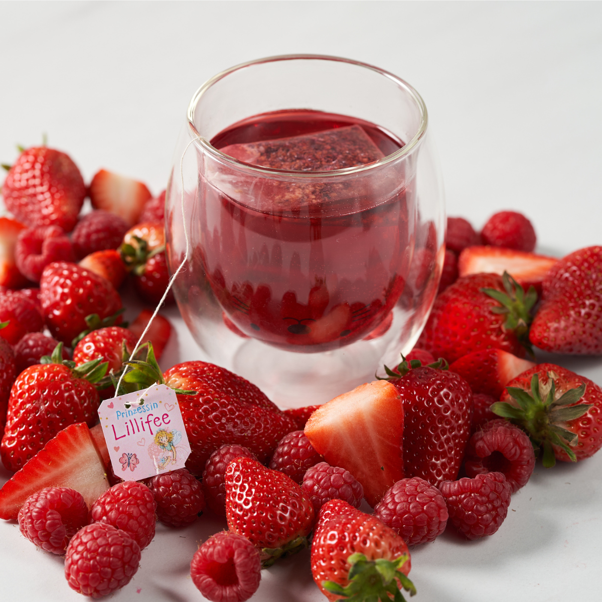 Eine Glastasse mit Prinzessin Lillifee-Tee steht auf einer weißen Fläche, umgeben von frischen Erdbeeren und Himbeeren, und zwischen den leuchtenden Beeren liegt ein Lillifee-Teebeutel.