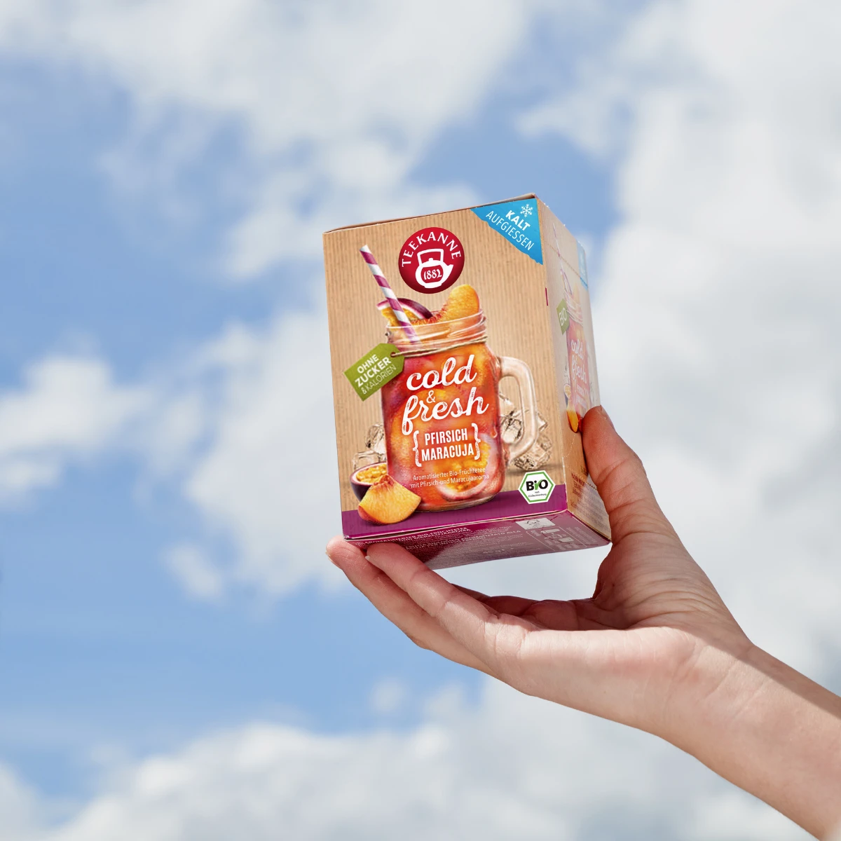 Eine Hand hält eine Schachtel kalten, frischen Mix & Match-Tee vor einem strahlenden, bewölkten Himmel hoch. Die Verpackung zeigt ein Einmachglas mit Tee, Fruchtscheiben und einem gestreiften Strohhalm – ideal für eine sommerliche Erfrischung.