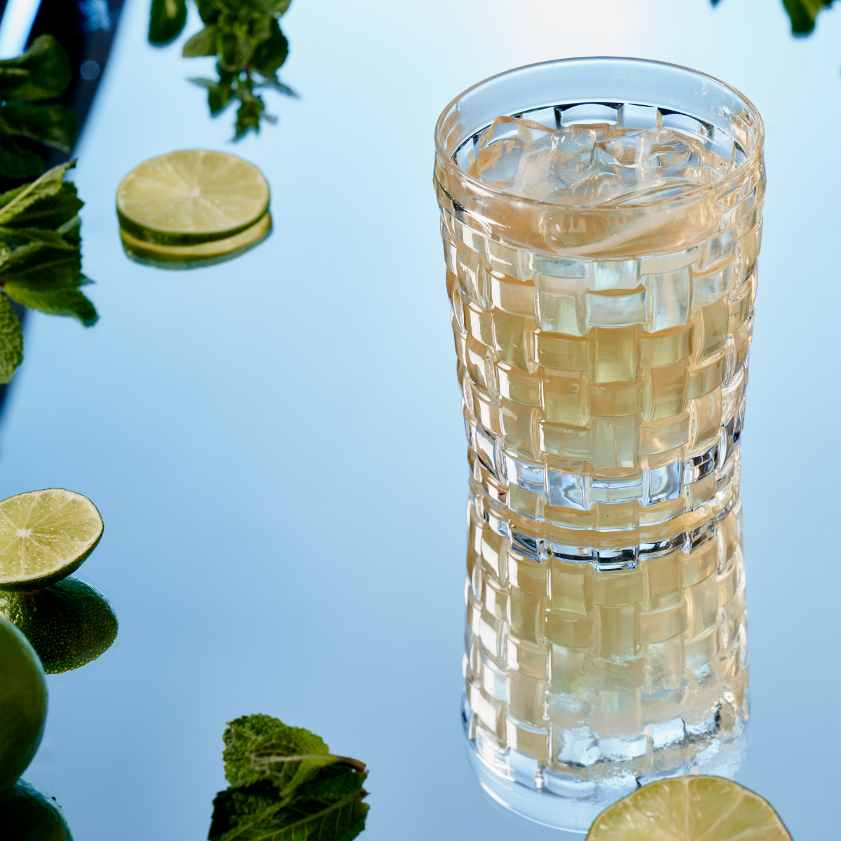 Ein Glas kaltes, frisches Mix & Match steht auf einer spiegelnden Oberfläche, umgeben von Minzblättern, Ingwer und Zitronenscheiben. Die Spiegelung des Getränks schimmert auf dem glänzenden Hintergrund.