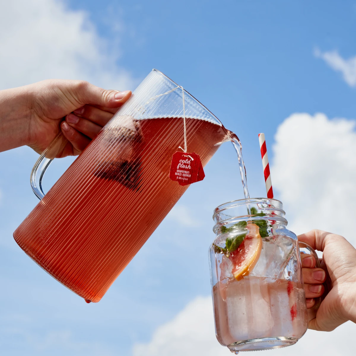 Eine Hand gießt vor einem blauen Himmel mit Wolken roten Eistee „Cold&Fresh + Frio Mix & Match“ aus einer Glaskanne mit Teebeuteletikett in ein Einmachglas mit Eis, Grapefruitscheiben und einem gestreiften Strohhalm.