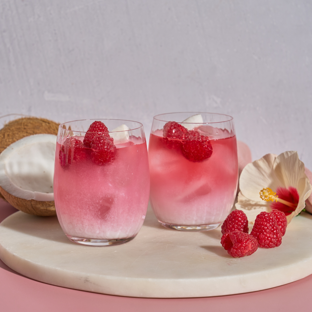 Zwei Cold&Fresh + Frio Mix & Match-Getränke aus rosa Kokosnuss und Himbeere in durchsichtigen Gläsern, jedes mit Himbeeren garniert, stehen auf weißem Marmor mit einer halbierten Kokosnuss, Himbeeren und einer rosa Blume in der Nähe.