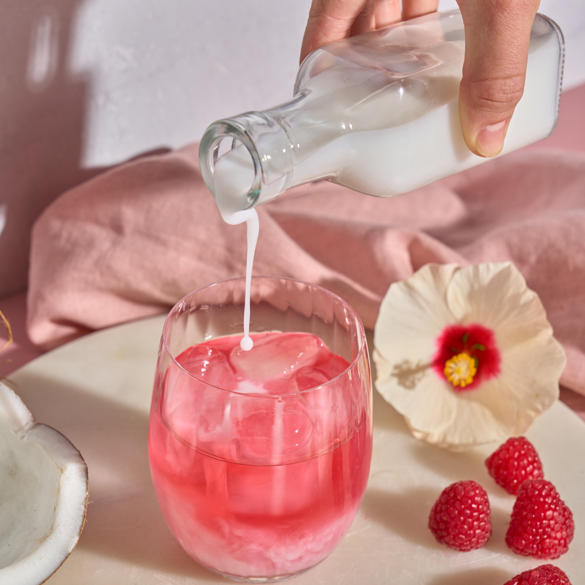 Eine Hand gießt eine cremeweiße Flüssigkeit in ein Glas mit rosa Frio Mix & Match Tee und Eis. Daneben liegen Himbeeren, eine Hibiskusblüte und eine Kokosnussschale auf einer hellen Oberfläche mit rosa Stoffhintergrund – ideal für Mix & Match-Momente.