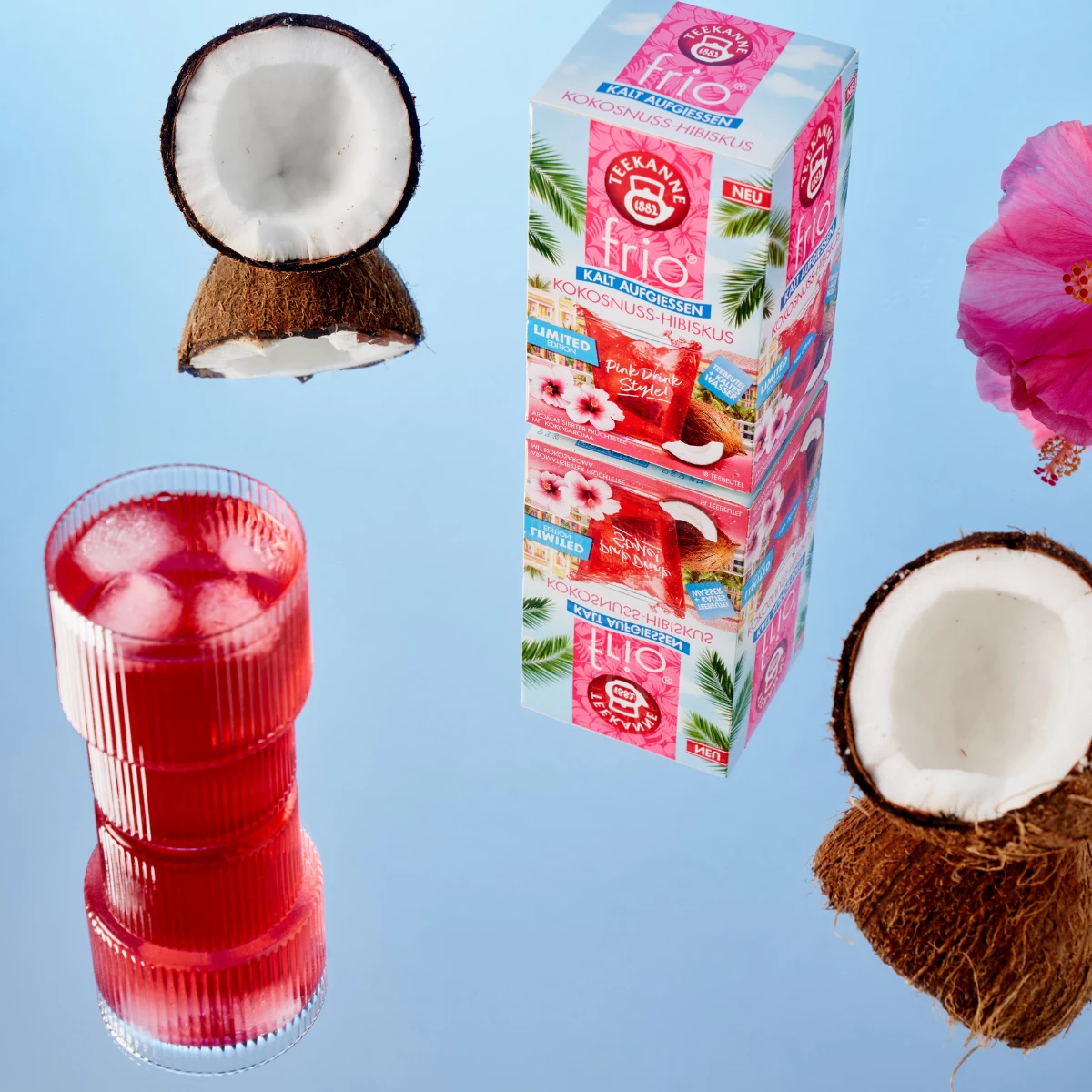 Eine Kokosnuss, eine rosa Hibiskusblüte, eine Schachtel Frio Mix & Match Kokos-Hibiskus-Tee und ein Glas leuchtend rosa Eistee werden auf einer reflektierenden Oberfläche mit blauem Hintergrund präsentiert – perfekt für den Genuss von kalt gebrühtem Tee.