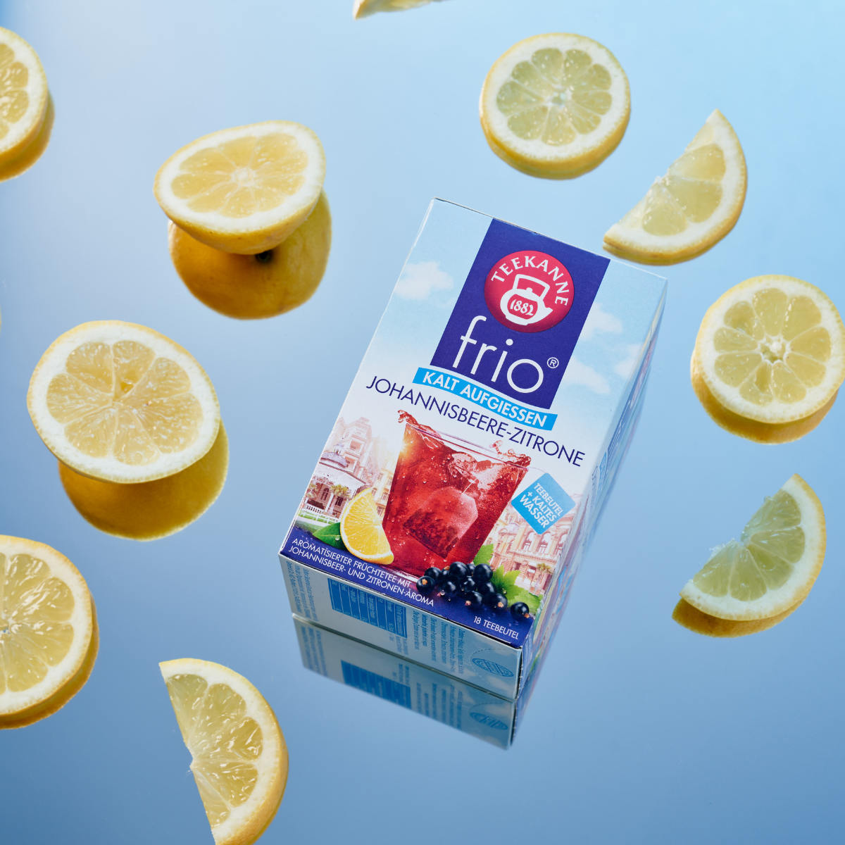 Eine Schachtel Frio Mix & Match Tee ist von Zitronenscheiben auf einer reflektierenden blauen Oberfläche umgeben. Die Verpackung präsentiert erfrischenden roten Eistee mit Zitrone und schwarzen Johannisbeeren, perfekt für die kalte Zubereitung mit Mix & M