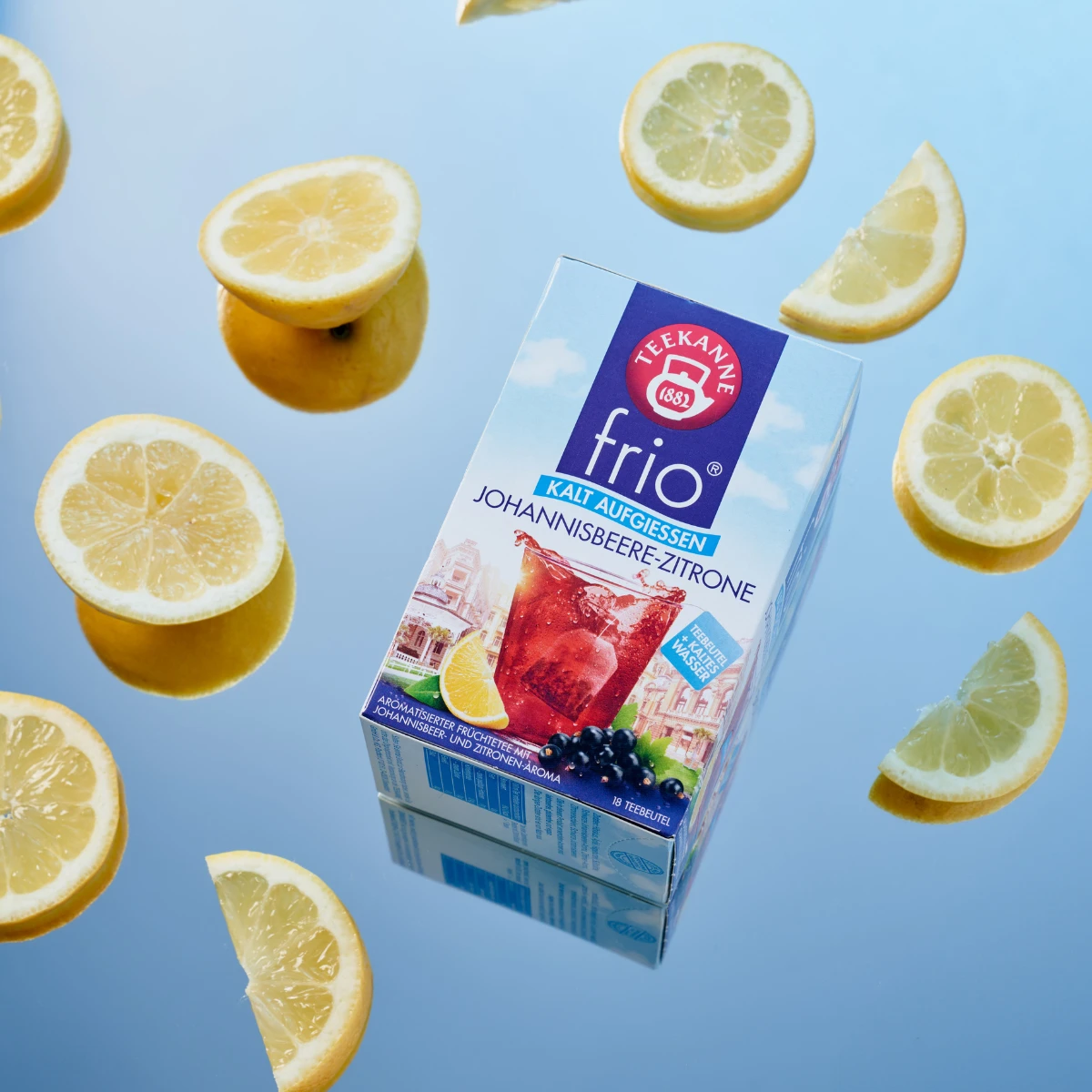 Eine Schachtel Frio Mix & Match Tee ist von Zitronenscheiben auf einer reflektierenden blauen Oberfläche umgeben. Die Verpackung präsentiert erfrischenden roten Eistee mit Zitrone und schwarzen Johannisbeeren, perfekt für die kalte Zubereitung mit Mix & M