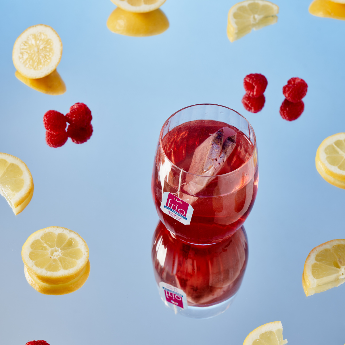 Ein Glas Frio Mix & Match Eistee mit einer Zimtstange, umgeben von Zitronenscheiben und Himbeeren, steht auf einer reflektierenden blauen Oberfläche.