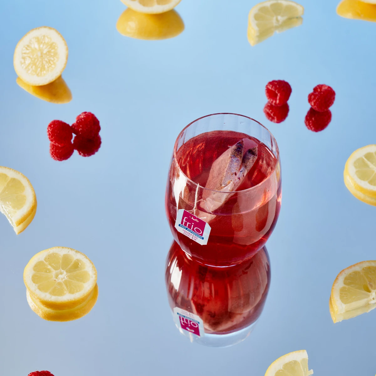 Ein Glas Frio Mix & Match Eistee mit einer Zimtstange, umgeben von Zitronenscheiben und Himbeeren, steht auf einer reflektierenden blauen Oberfläche.