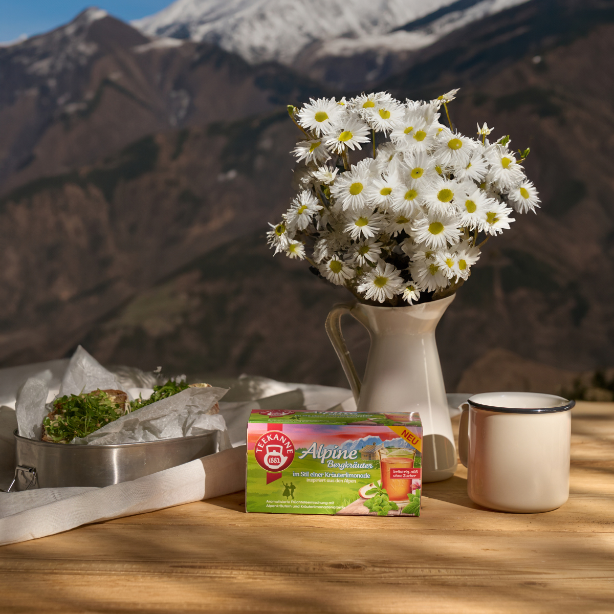 Ein Strauß Gänseblümchen, eine weiße Tasse, eine TEEKANNE-Teebox und Sandwiches auf einem Holztisch laden dazu ein, Ländertee Mix & Match Früchtetees im Freien mit Bergen im Hintergrund zu genießen.