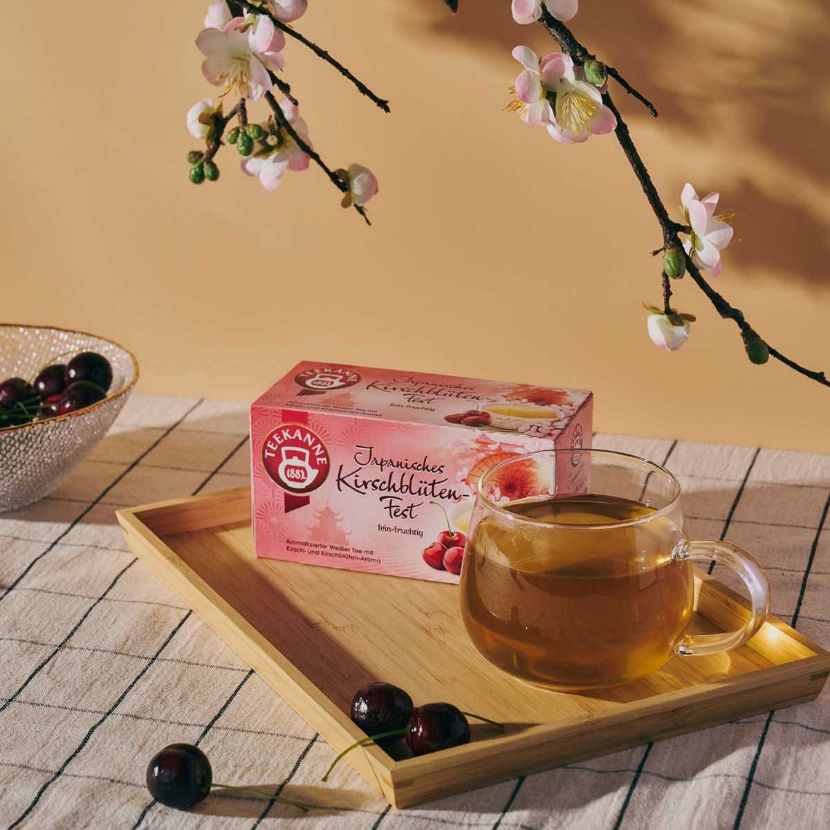 Auf einem Bambustablett steht eine Teetasse aus Glas neben einer Schachtel Ländertee Mix & Match. Kirschblüten und Kirschen sind daneben auf einer karierten Tischdecke mit pfirsichfarbenem Hintergrund arrangiert.