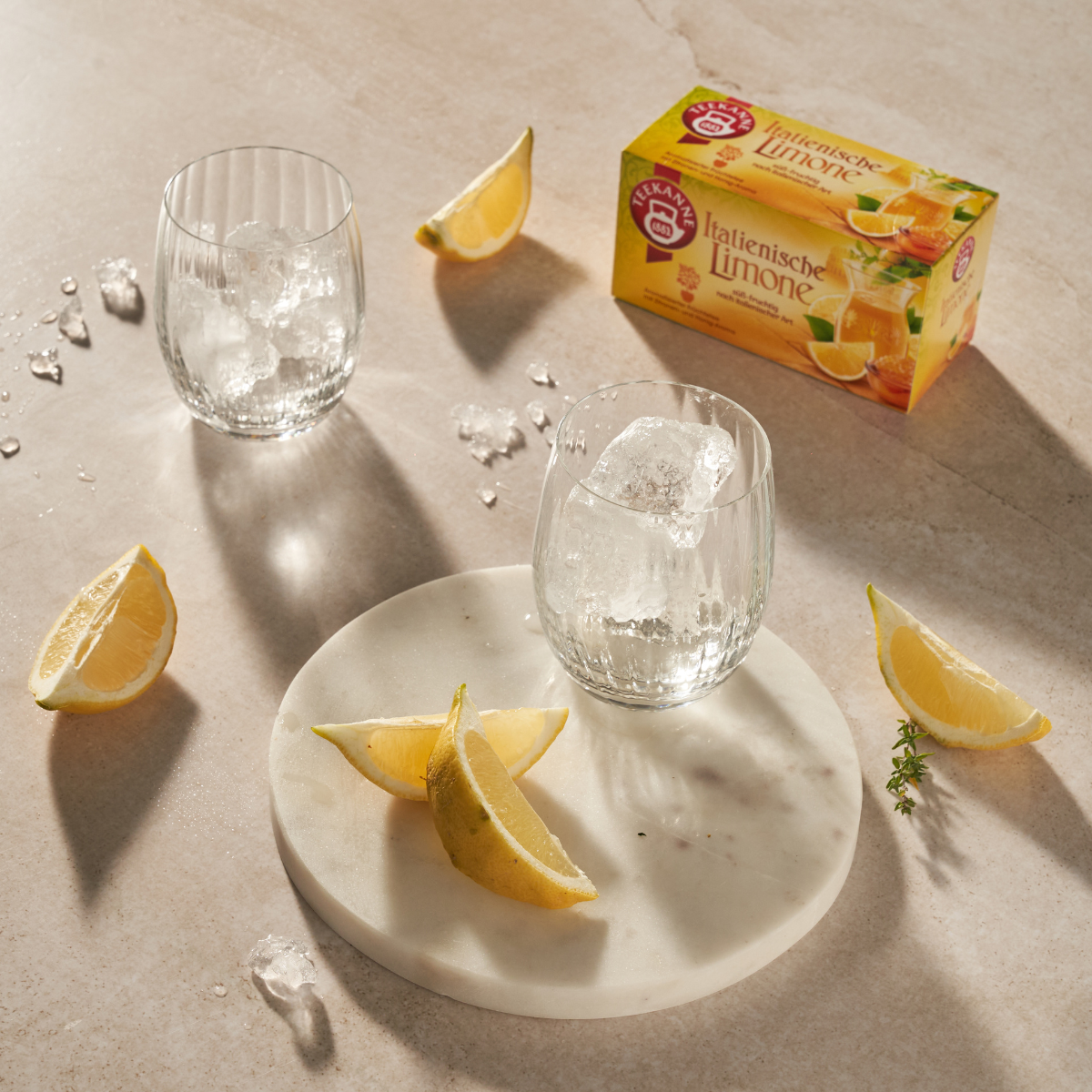 Zwei Gläser mit Eis, Zitronenspalten und verstreuten Eiswürfeln stehen auf einer hellen Fläche neben einem Marmortablett mit Zitronenscheiben. Im Hintergrund fängt eine Ländertee Mix & Match Box sanftes Sonnenlicht ein und wirft sanfte Schatten.