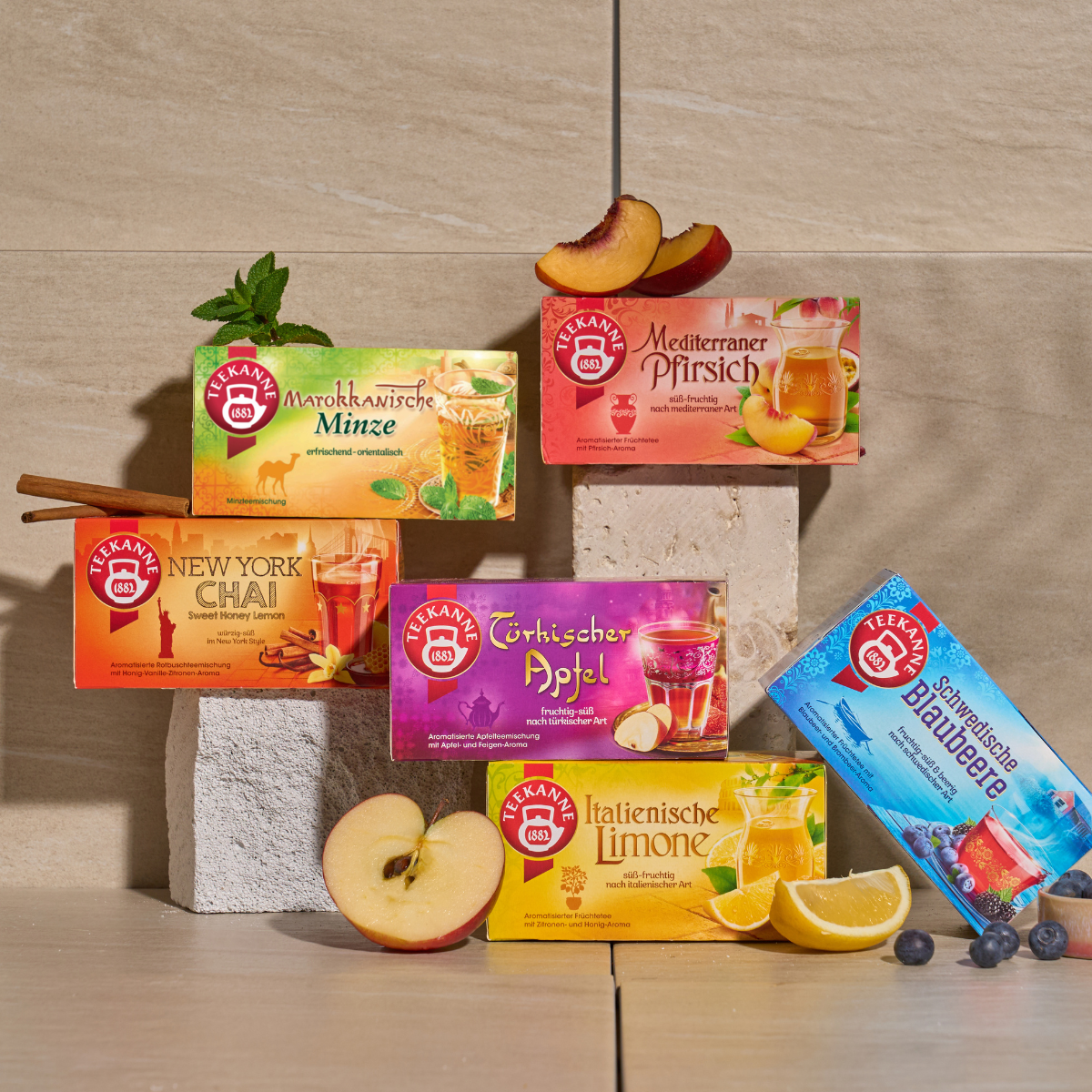 Sechs Schachteln Ländertee Mix & Match Tees in verschiedenen Geschmacksrichtungen – Apfel, Pfirsich, Zitrone, Blaubeere und Pflaume – werden auf Steinblöcken mit passenden Früchten präsentiert. Der neutrale beige Fliesenhintergrund hebt die Vielfalt hervo
