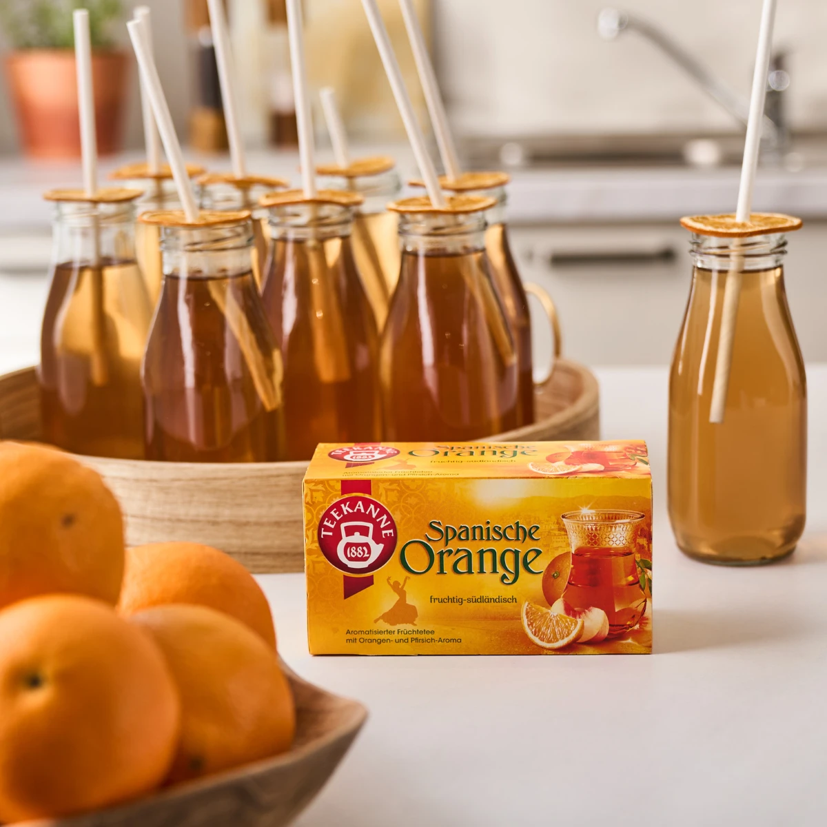 Eine Schachtel Ländertee Mix & Match steht auf einer Küchentheke, umgeben von orangefarbenen Teeflaschen mit Strohhalmen und Orangenscheiben. Frische Orangen und die farbenfrohe Verpackung runden das Bild ab.