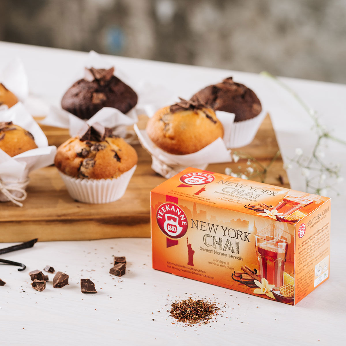 Eine Schachtel New York Chai-Tee steht auf einem weißen Tisch neben Schokoladen- und Vanillemuffins auf einem Holztablett, umgeben von Schokoladenstückchen, Honigtropfen und losem Rooibos-Tee.