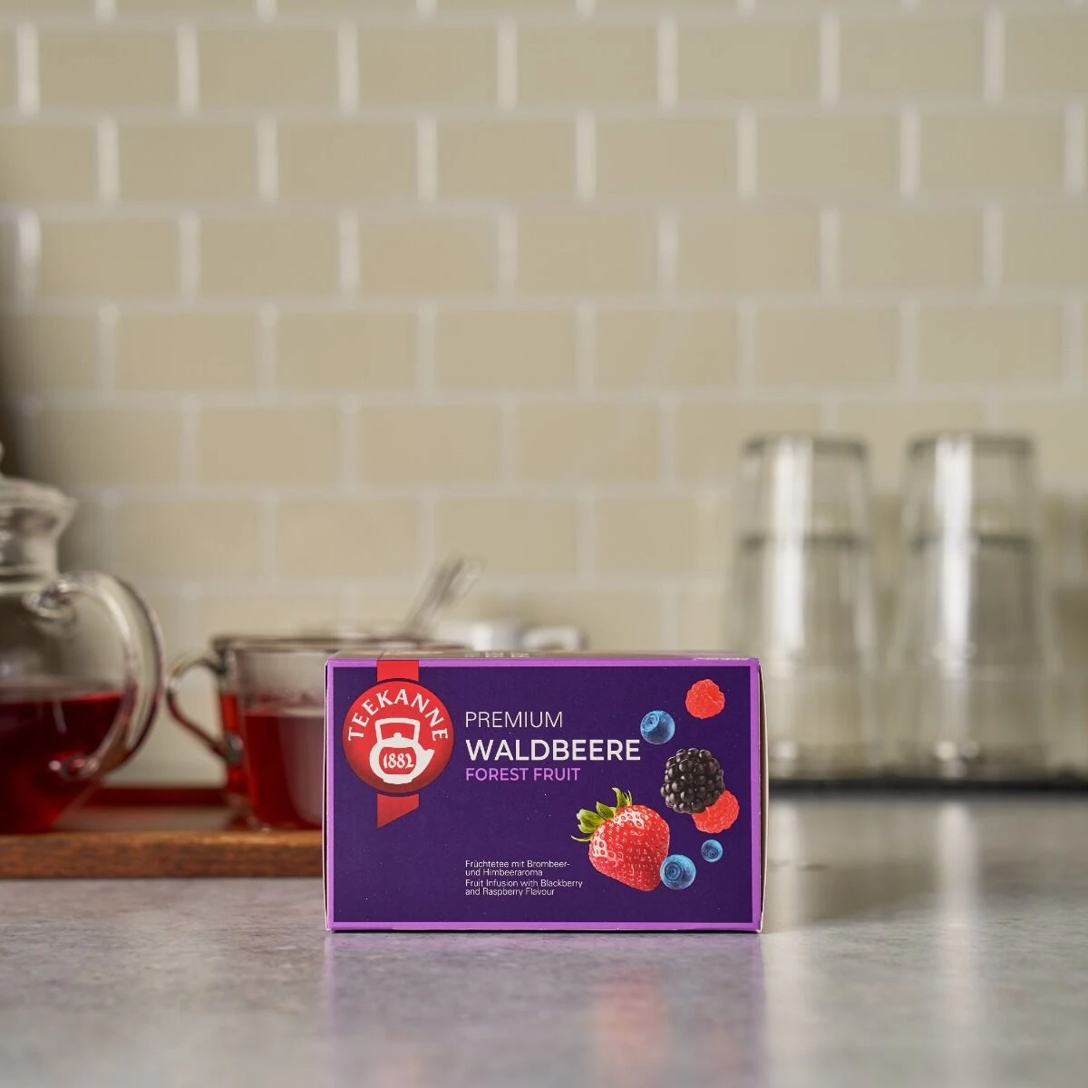 Auf einem Küchentisch steht eine Schachtel Premium Waldbeeren-Tee mit einer Teekanne und Glastassen. Die Verpackung ist mit bunten Beerenmotiven verziert und steht auf einer gefliesten Arbeitsfläche.