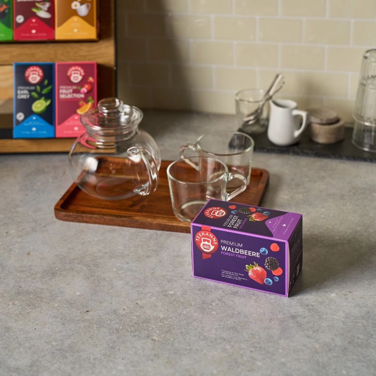 Eine Schachtel Premium Waldbeeren-Tee steht auf einem Küchentisch neben einer gläsernen Teekanne, zwei durchsichtigen Tassen auf einem Holztablett und mehreren anderen Teeschachteln, die in einem Regal im Hintergrund gestapelt sind.