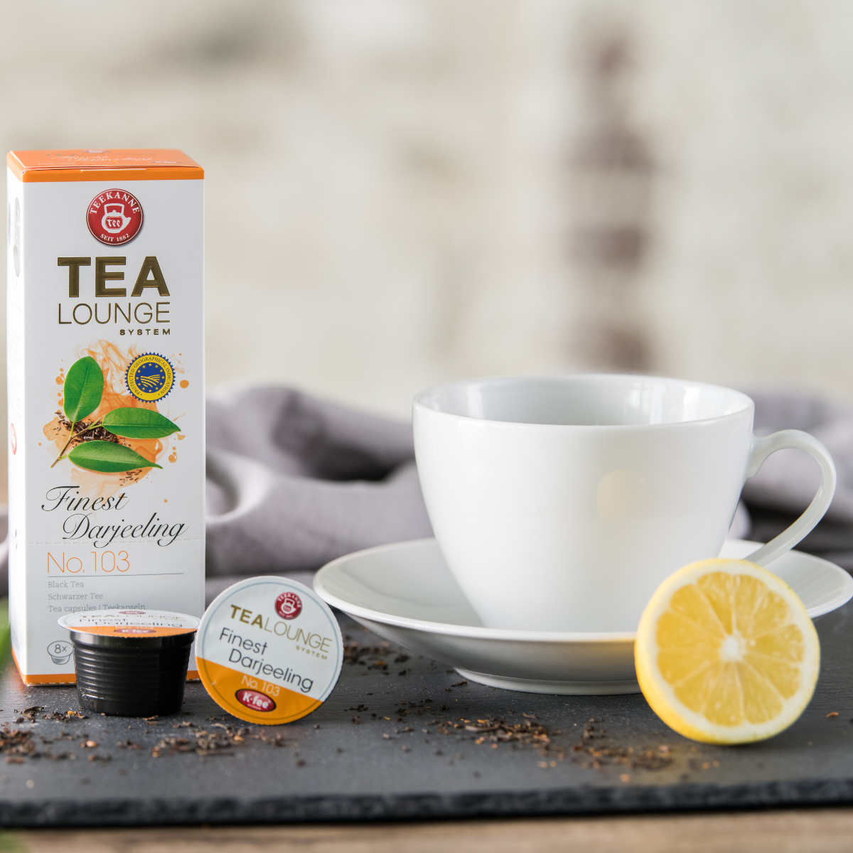 Eine weiße Teetasse auf einer Untertasse steht neben einer Schachtel Finest Darjeeling Nr. 103, einer Teekapsel, einer schwarzen Teekapsel, losen Schwarzteeblättern und einer Zitronenscheibe auf einer dunklen Oberfläche mit einem grauen Tuch im Hintergrun