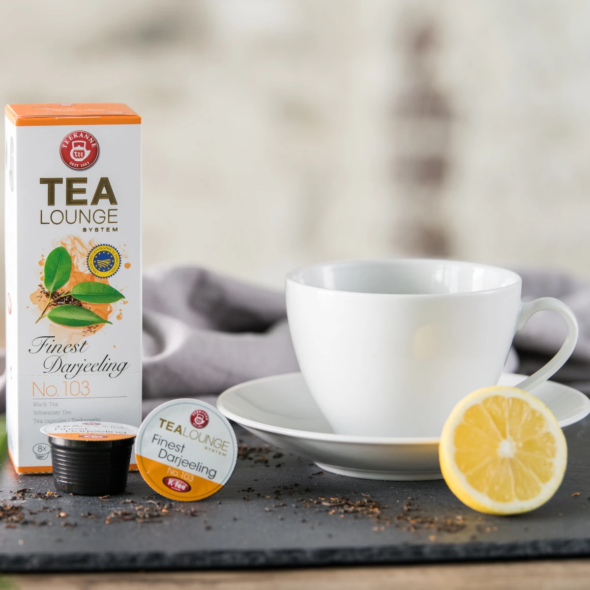 Eine weiße Teetasse auf einer Untertasse steht neben einer Schachtel Finest Darjeeling Nr. 103, einer Teekapsel, einer schwarzen Teekapsel, losen Schwarzteeblättern und einer Zitronenscheibe auf einer dunklen Oberfläche mit einem grauen Tuch im Hintergrun