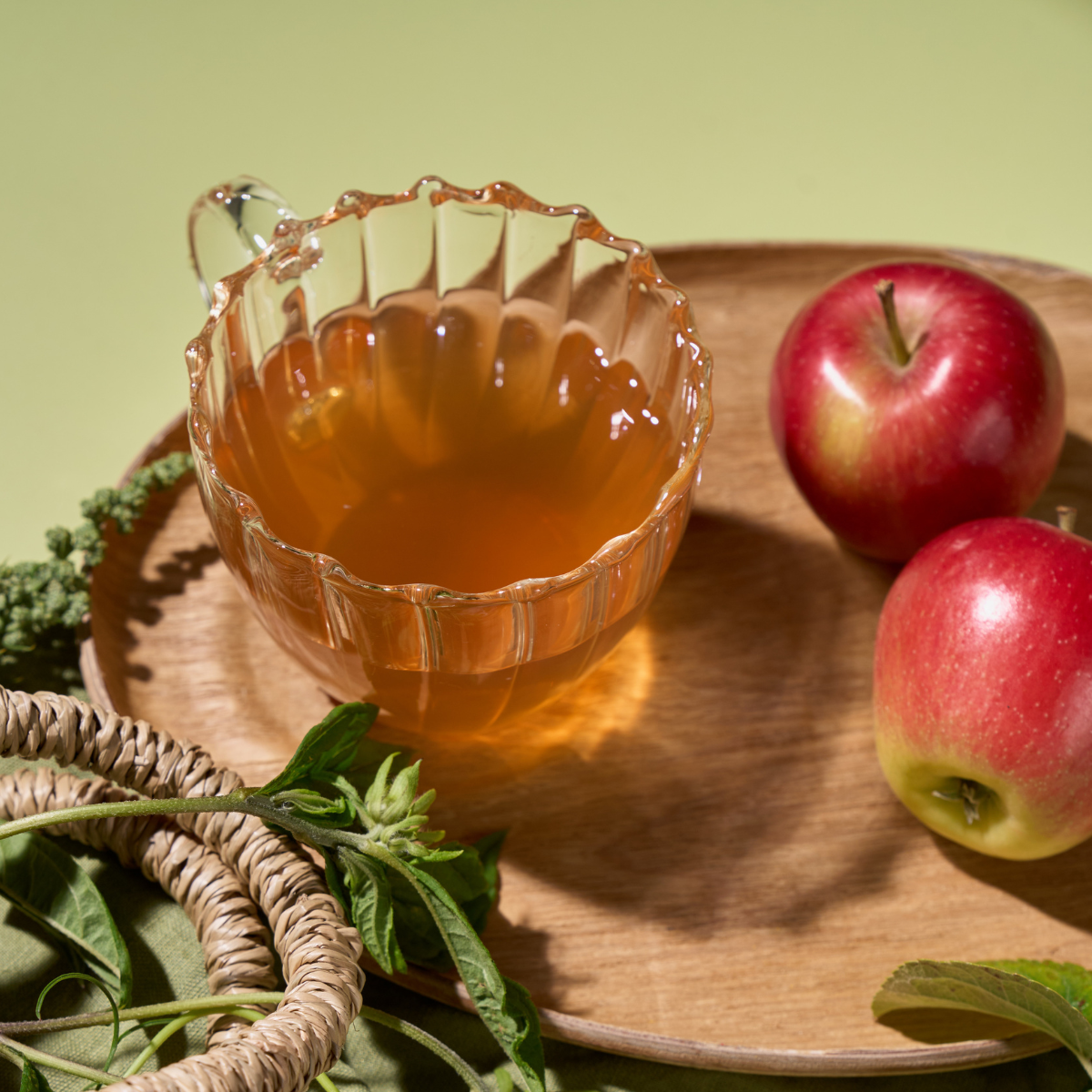 Eine Glastasse mit Bio Apfel Holunderblüte Tee steht auf einem Holzteller neben zwei roten Äpfeln, grünen Kräutern und Weidenringen und lädt zu einem fruchtigen Genussgarten-Moment vor einem sanften grünen Hintergrund ein.