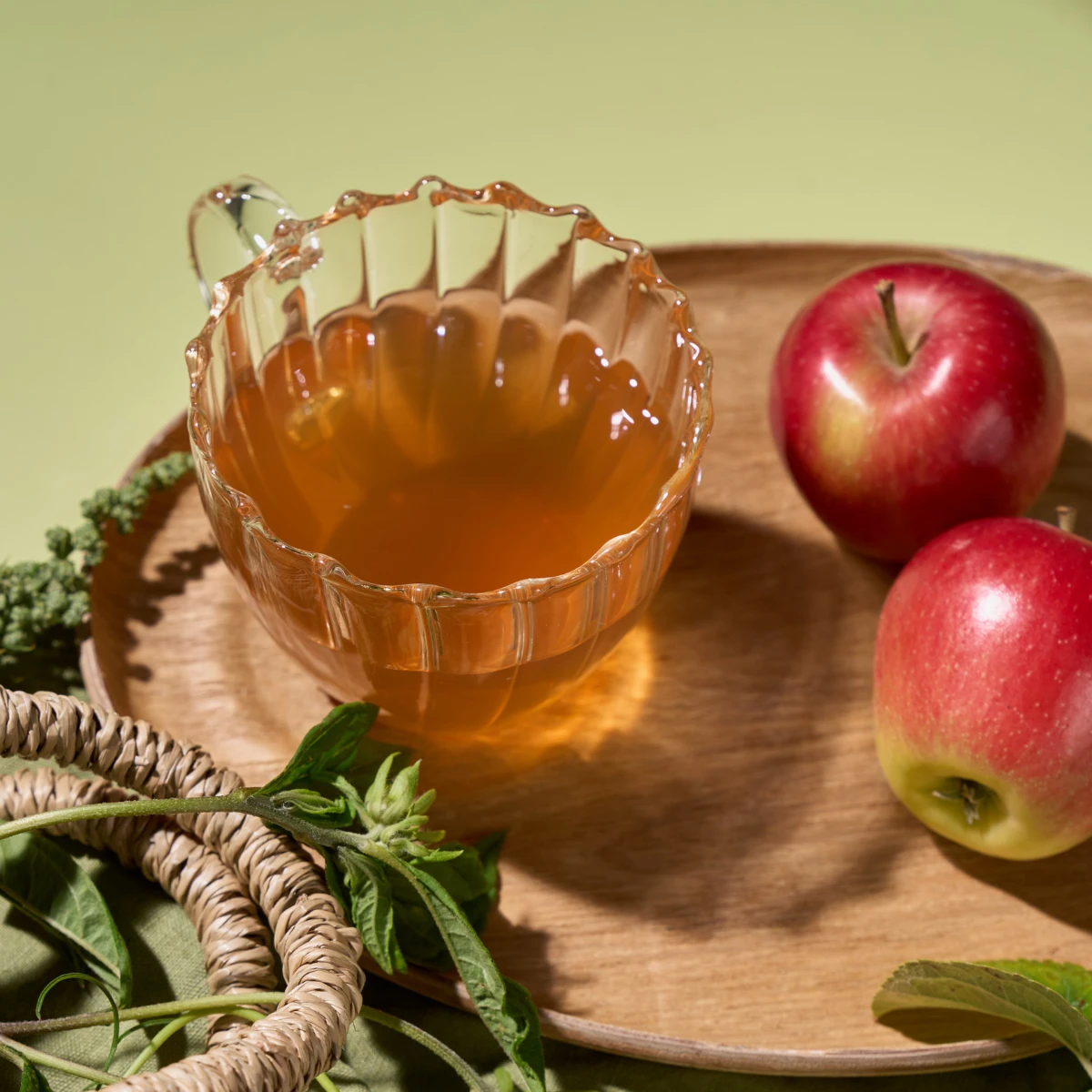 Eine Glastasse mit Bio Apfel Holunderblüte Tee steht auf einem Holzteller neben zwei roten Äpfeln, grünen Kräutern und Weidenringen und lädt zu einem fruchtigen Genussgarten-Moment vor einem sanften grünen Hintergrund ein.