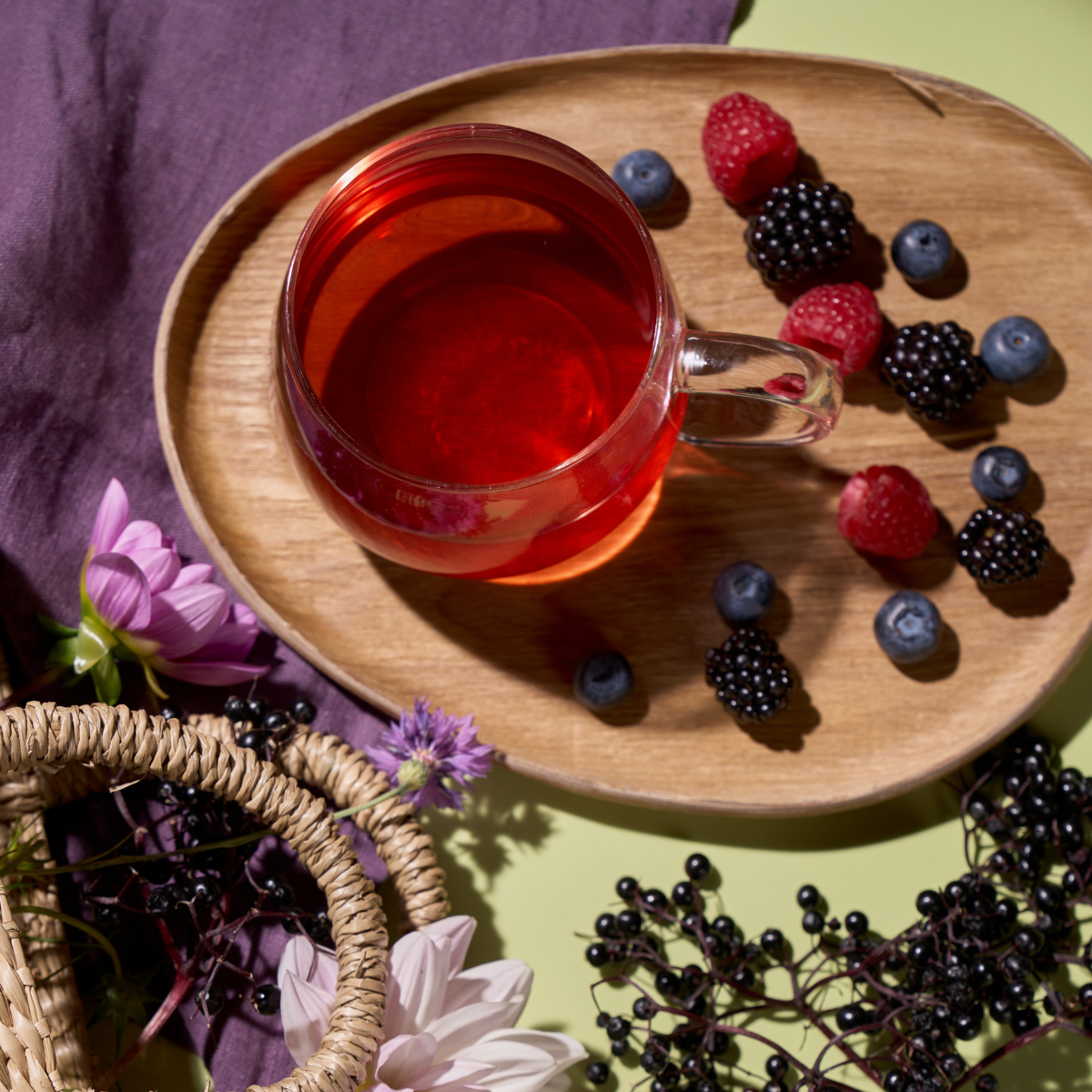 Eine Glastasse mit rotem Tee auf einem Holzteller, umgeben von Himbeeren, Brombeeren, Heidelbeeren, violetten Blumen und einem geflochtenen Korb auf einer hellgrünen Fläche mit einem violetten Tuch - reiner Bio Holunder Waldbeere Genuss.