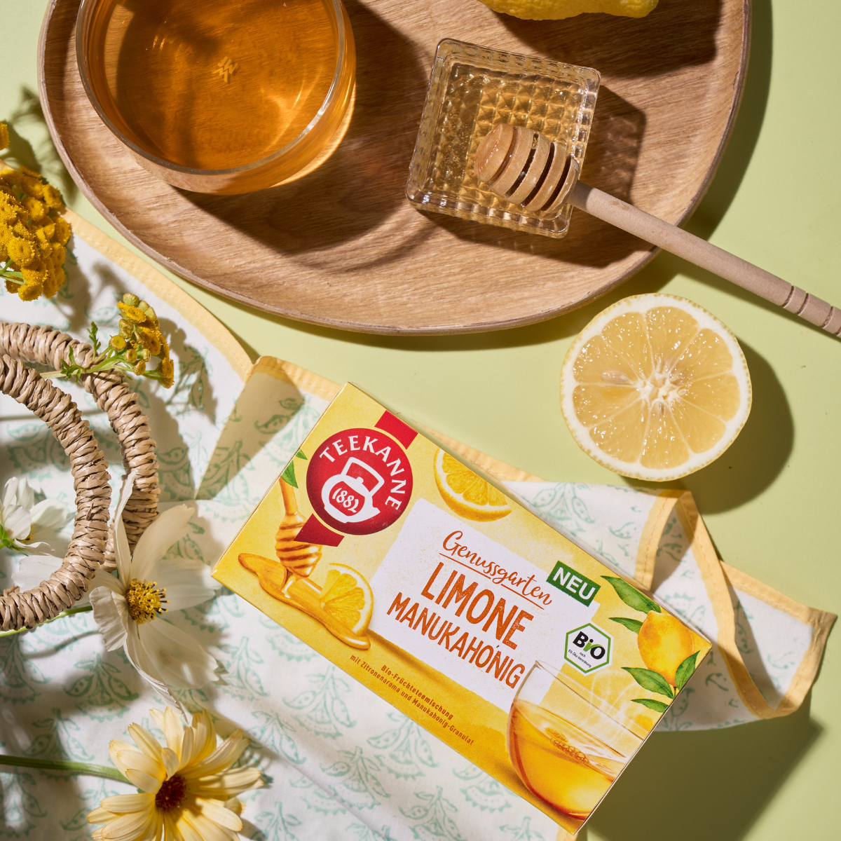 Eine Schachtel Bio Limone Manukahonig-Tee steht auf einem Tisch mit einer halbierten Zitrone, einem Honiglöffel in einem Glas, einer Tasse Früchtetee, gelben Blumen und einem Blumentuch, alles in hellem Tageslicht arrangiert.