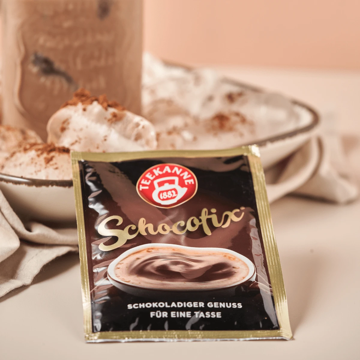 Ein Schocofix Beutel 25g liegt vor einer Tasse heißer Schokolade mit Schlagsahne, während im Hintergrund Kakaopulver auf den Teller gestreut ist.