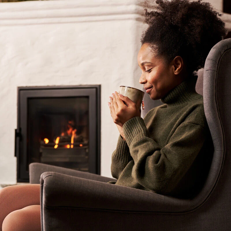 Eine Frau sitzt lächelnd mit geschlossenen Augen in einem grauen Sessel, hält eine Tasse Wintertee Mix & Match in der Hand und entspannt in gemütlicher Atmosphäre vor einem brennenden Kaminfeuer.