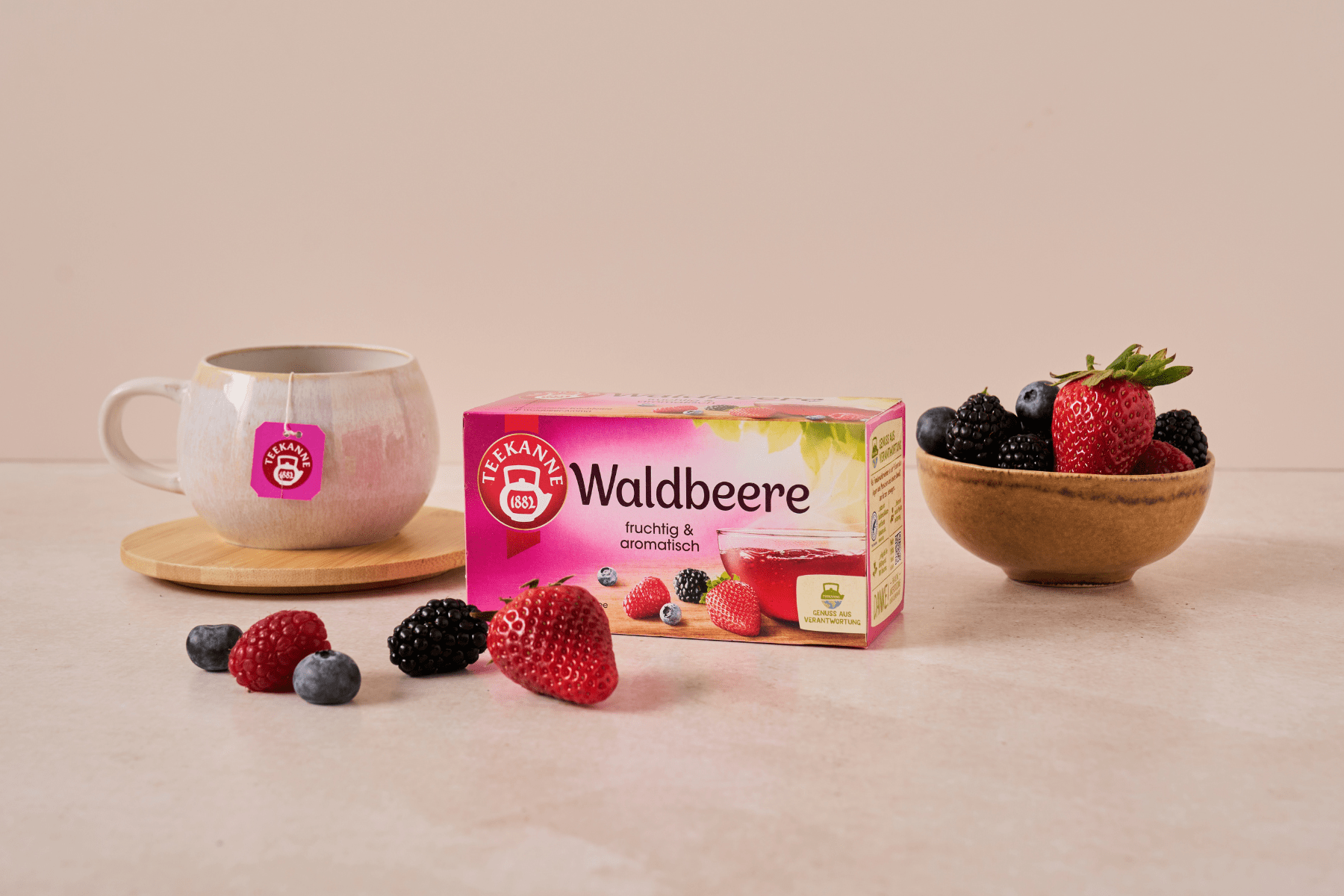 Eine Schachtel Kräuter- und Früchtetee Mix & Match steht neben einer Teetasse auf einer hölzernen Untertasse und einer Schale mit gemischten Beeren, wie Erdbeeren, Blaubeeren, Himbeeren und Brombeeren.