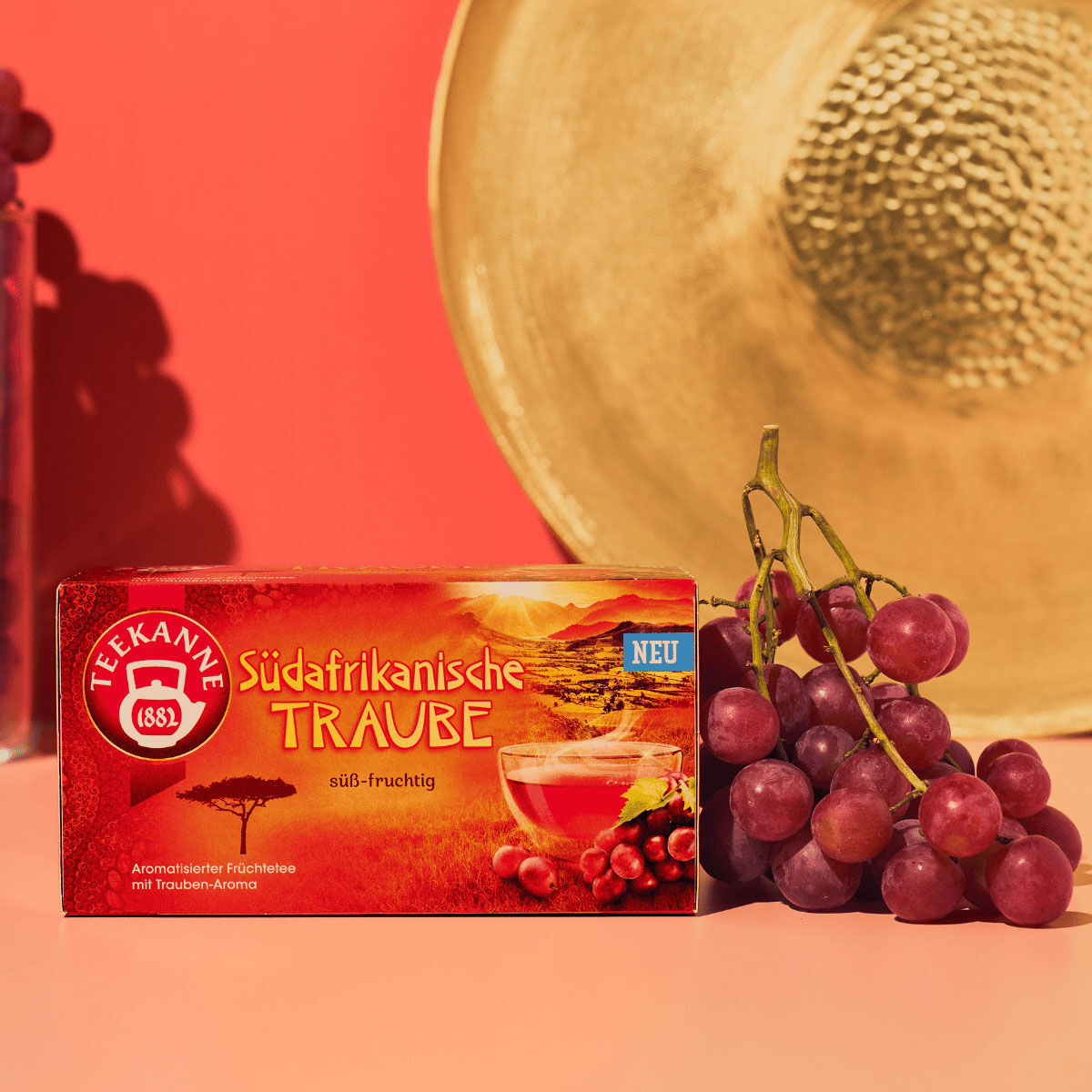 Eine Schachtel südafrikanischer Traube-Tee steht neben roten Trauben auf einer rosa Oberfläche, im Hintergrund sind ein Strohhut und ein Glas Trauben zu sehen. Die Szene ist hell und farbenfroh.
