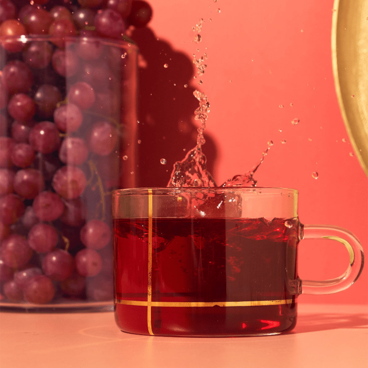 Eine Tasse aus klarem Glas, in der roter Tee spritzt, steht vor einem Glas mit der Aufschrift „Südafrikanische Traube“, vor einem leuchtend rosa und roten Hintergrund.