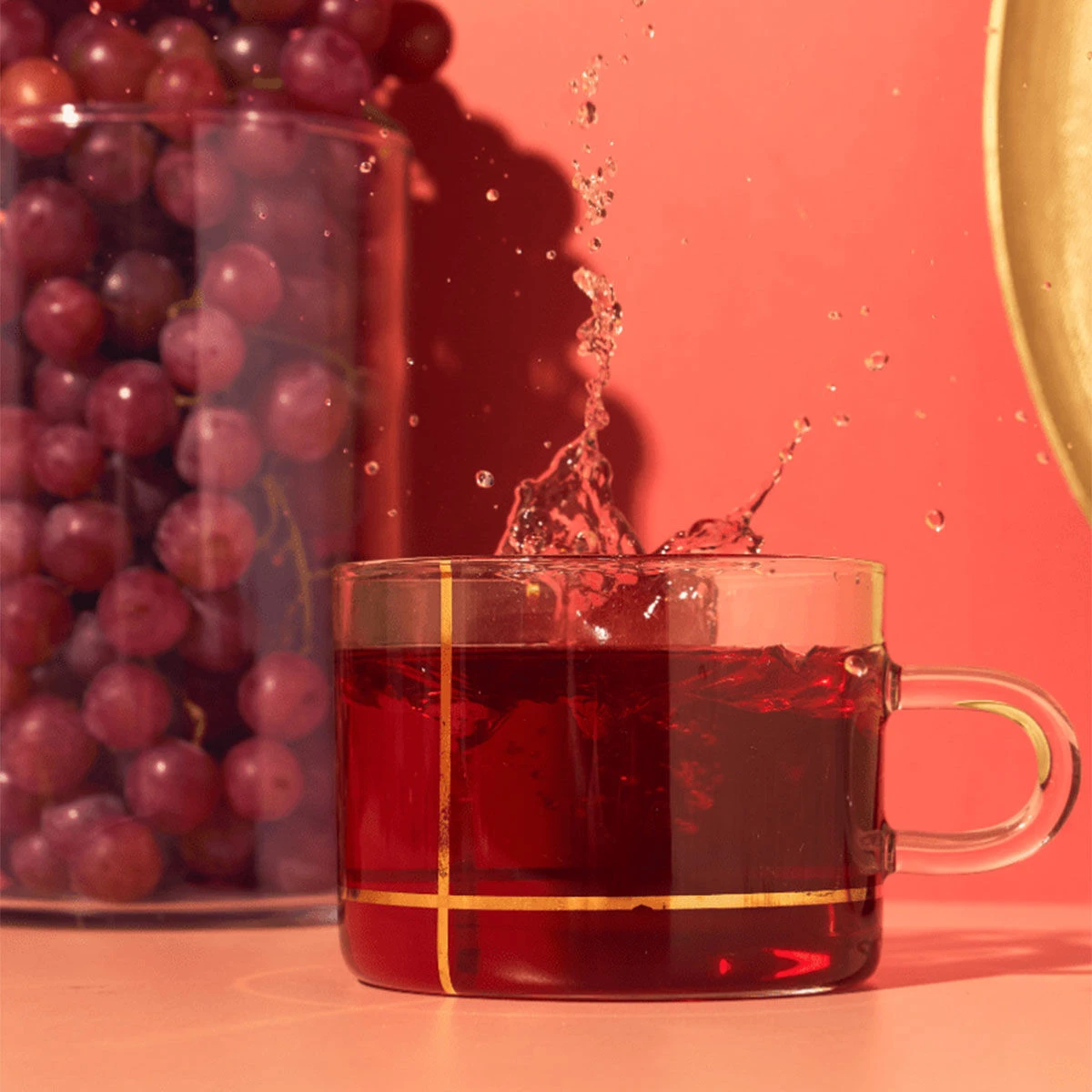 Eine Tasse aus klarem Glas, in der roter Tee spritzt, steht vor einem Glas mit der Aufschrift „Südafrikanische Traube“, vor einem leuchtend rosa und roten Hintergrund.