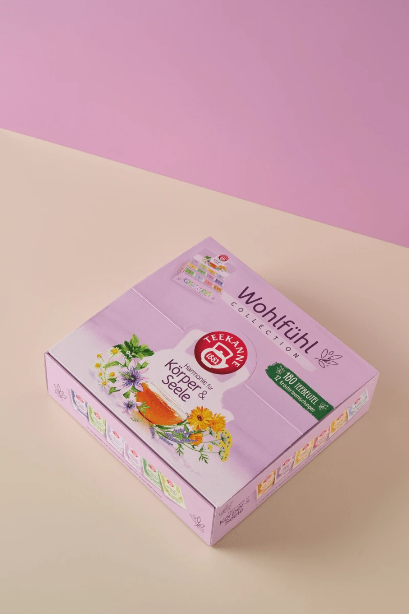 Auf einer pastellfarbenen Oberfläche sitzt eine rosafarbene Wohlfühl-Collection Box Geschenkset - 180 Beutel, die mit Kräutern und Blumen sowie dem roten Teekanne-Logo verziert ist und mit jeder Tasse zum Wohlfühlen & Genießen einlädt.