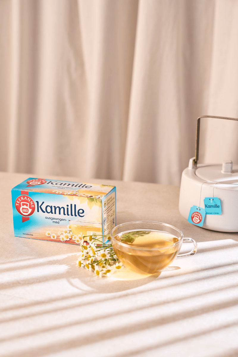 Eine Schachtel Kräuter- und Früchtetee Mix & Match, eine Glastasse mit Blütentee und ein Teezubereiter auf einem sonnenbeschienenen Tisch bilden den perfekten Rahmen, um Ihre Lieblingskräutermischungen zu genießen.