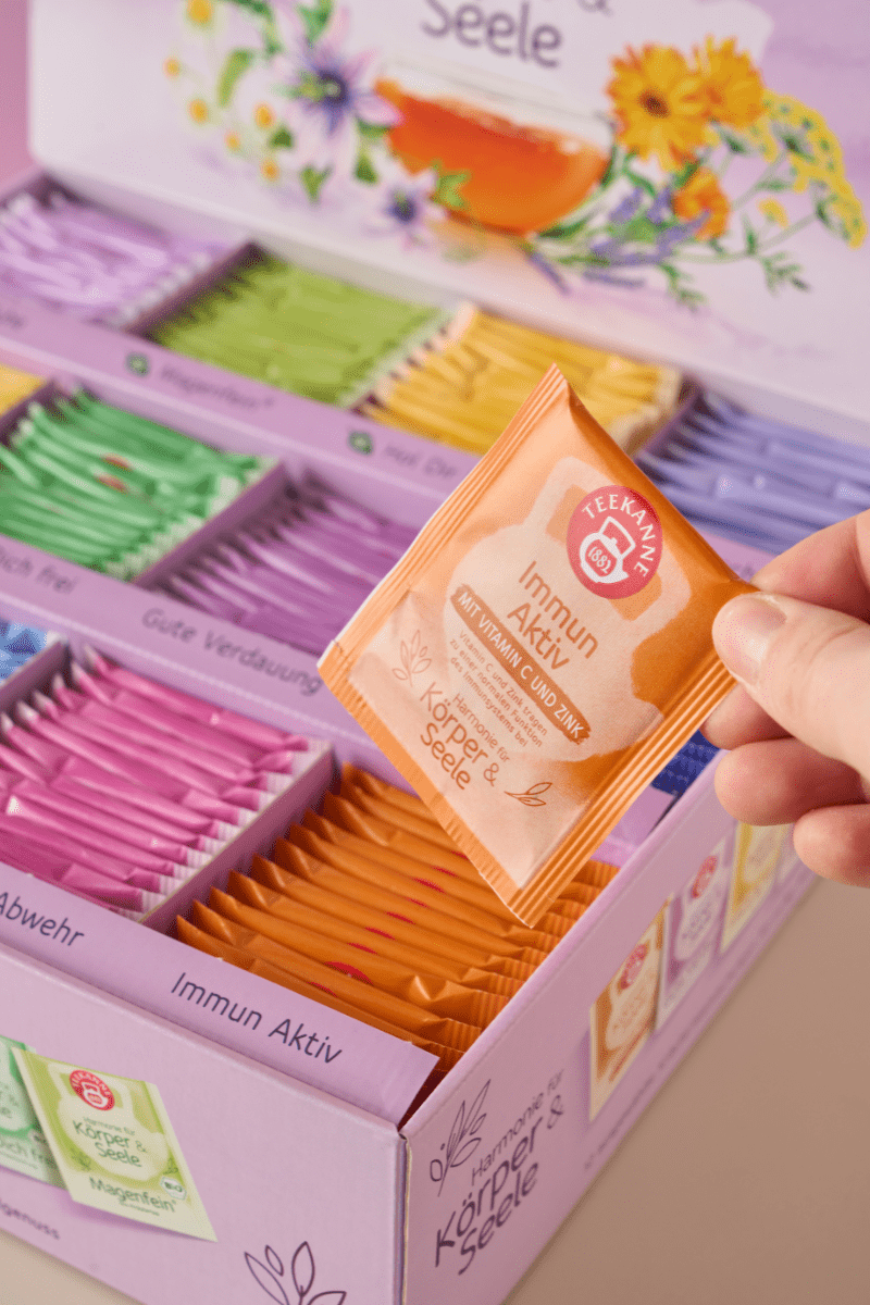 Eine Hand hält eine orangefarbene Immun Aktiv-Teepackung über der Teekanne Wohlfühl-Collection Box 180er, die verschiedene, farbenfrohe Kräuterteemischungen mit floralen Motiven und deutschem Text auf der lebendigen Verpackung enthält.