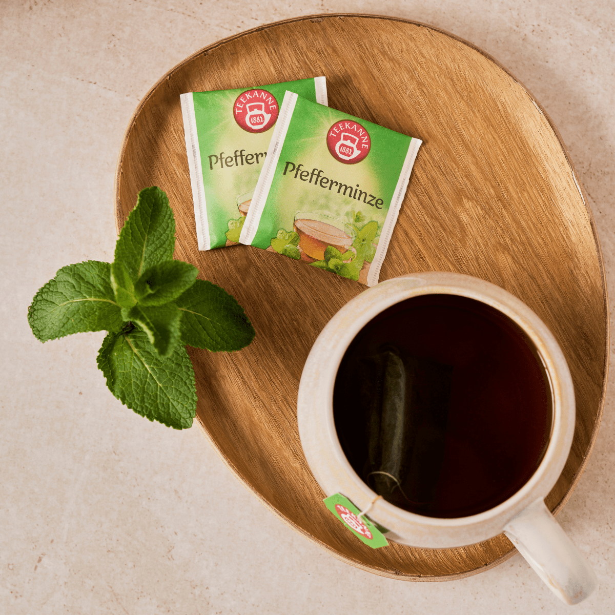 Ein Holztablett mit einer Tasse Pfefferminztee, zwei Tütchen Kräuter- und Früchtetee Mix & Match und einem Zweig frischer Minze auf einer hellen Unterlage.
