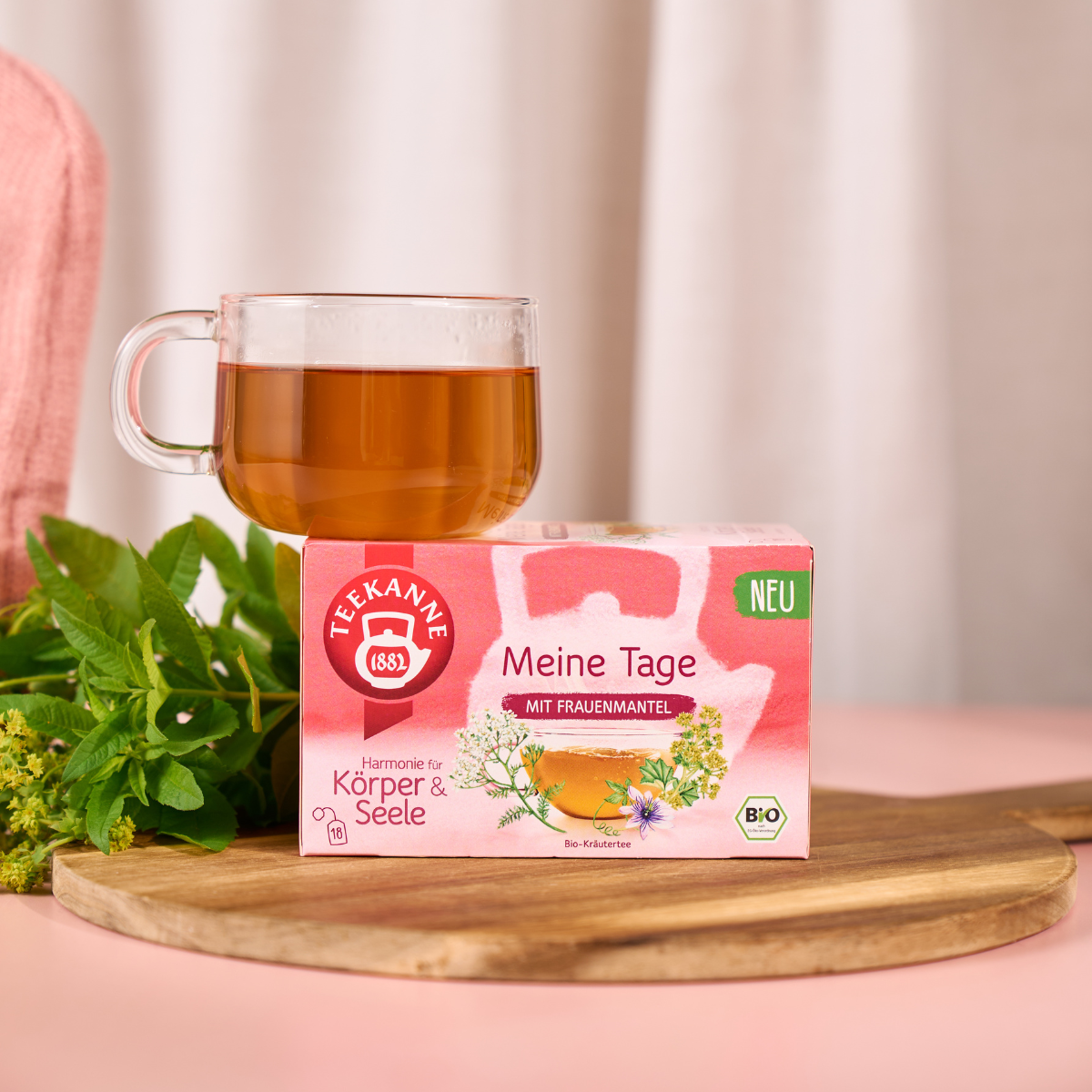 Eine Glastasse Kräutertee steht auf einem Holzbrett neben einer rosafarbenen Meine Tage-Teebox, frischen Frauenmantel- und Salbei-Kräutern und Blumen. Im sanft verschwommenen Hintergrund sind ein rosa Tuch und ein Vorhang zu sehen.