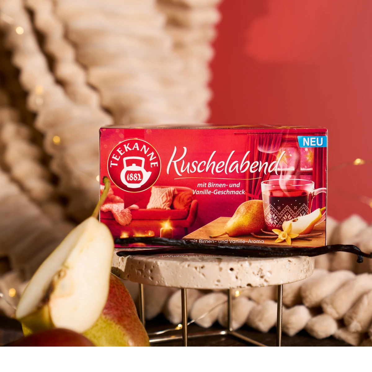 Eine Schachtel Kuschelabend-Tee ruht auf einem Marmorständer, begleitet von einer Birnenscheibe und einer Vanilleschote. Gemütliche Rot- und Beigetöne und verschwommenes Licht schaffen eine warme Atmosphäre.