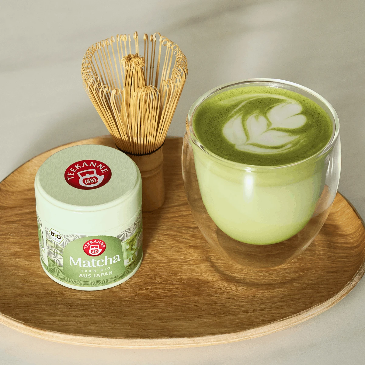 Ein Holztablett mit einer Dose Bio-Matcha, einem Bambusbesen und einem Glas Matcha-Latte mit blattförmiger Latte-Art.