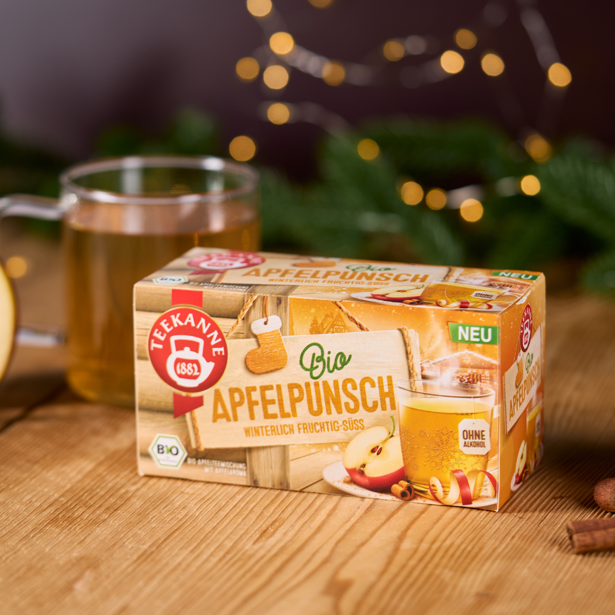 Eine Schachtel Bio Apfelpunsch steht auf einem Holztisch neben einer Glastasse mit Tee. Im Hintergrund leuchten festliche Lichter und Grünzeug, und auf der Verpackung sorgen Äpfel und Zimt für gemütlichen Wintergenuss.