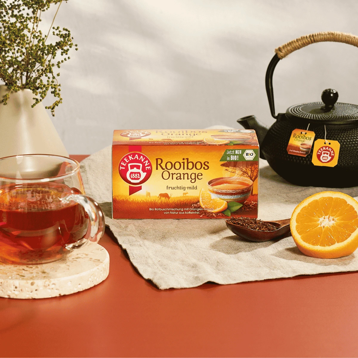 Auf einem roten Tisch steht eine Schachtel „Bio Rooibos Orange“-Tee auf einem Tischtuch, daneben eine Teekanne, eine Glas-Tasse mit mildem, fruchtigem Tee, eine in Scheiben geschnittene Orange, lose Teeblätter und eine Vase mit Blumen.