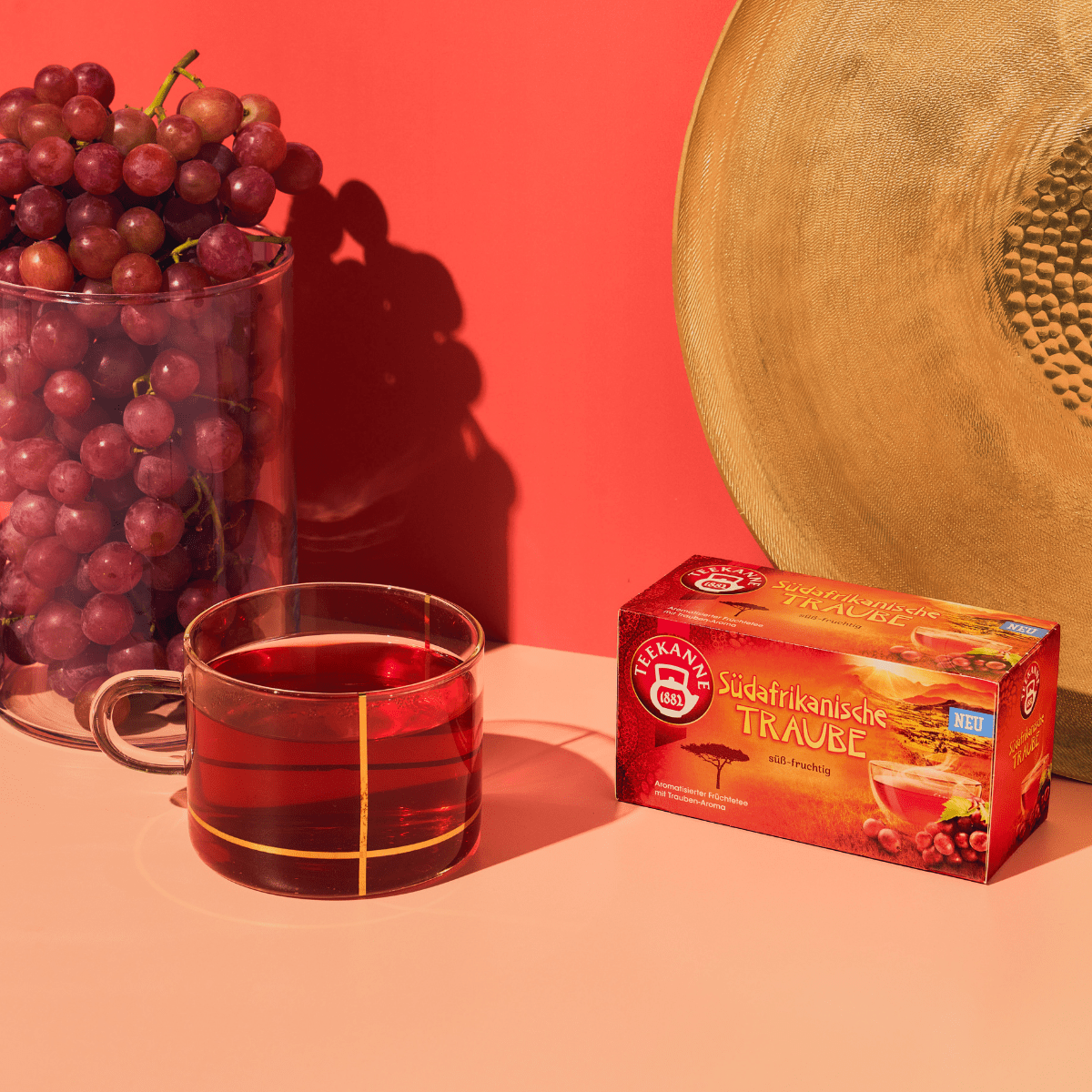 Eine Packung TEEKANNE Tee "Südafrikanische Traube" vor rötlichem Hintergrund und beigem Untergrund. Daneben eine gläserne Tasse mit aufgeossenem, rötlichem Tee. Dazu dekoriert mit roten Trauben und einer runden, goldenen Schale im Hintergrund.