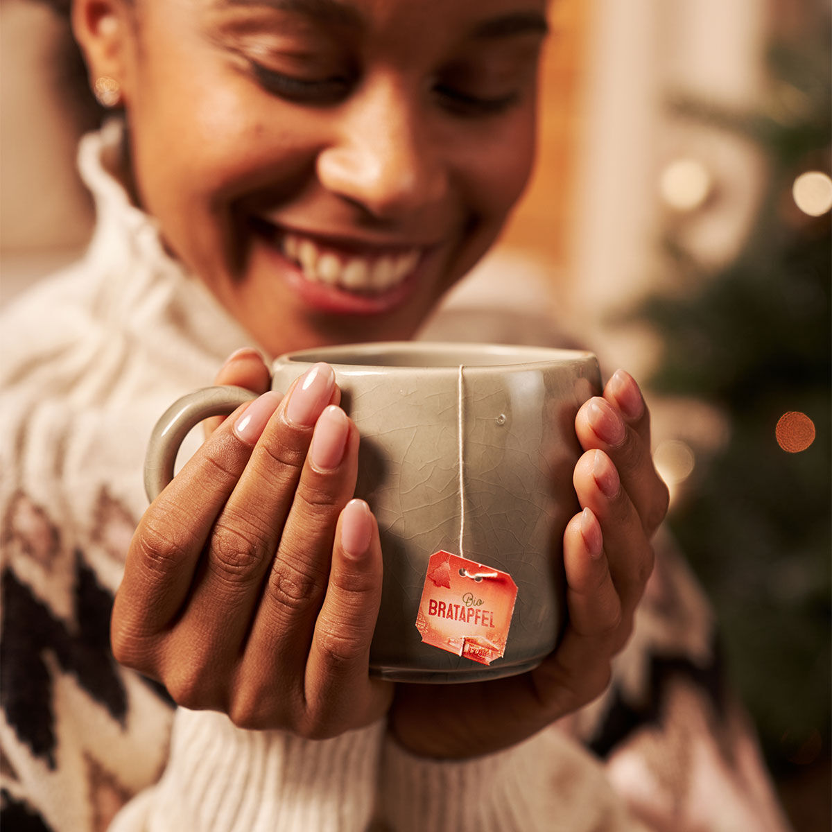 Eine Frau lächelt herzlich und hält eine große graue Tasse Bio-Bratapfel-Tee in der Hand. In einem kuscheligen gemusterten Pullover sitzt sie neben einem verschwommenen Weihnachtsbaum. Das rote Bio-Bratapfel-Etikett verleiht ihr festlichen Charme.
