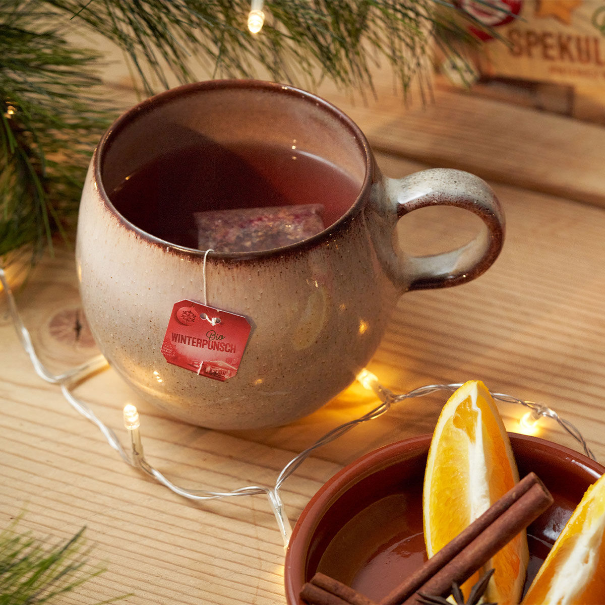 Eine Keramiktasse Bio-Winterpunsch-Tee mit einem „Winterpunsch“-Teebeutel steht auf einem Holztisch, umgeben von Tannennadeln, Lichterketten, getrockneten Orangenscheiben und Zimtstangen.