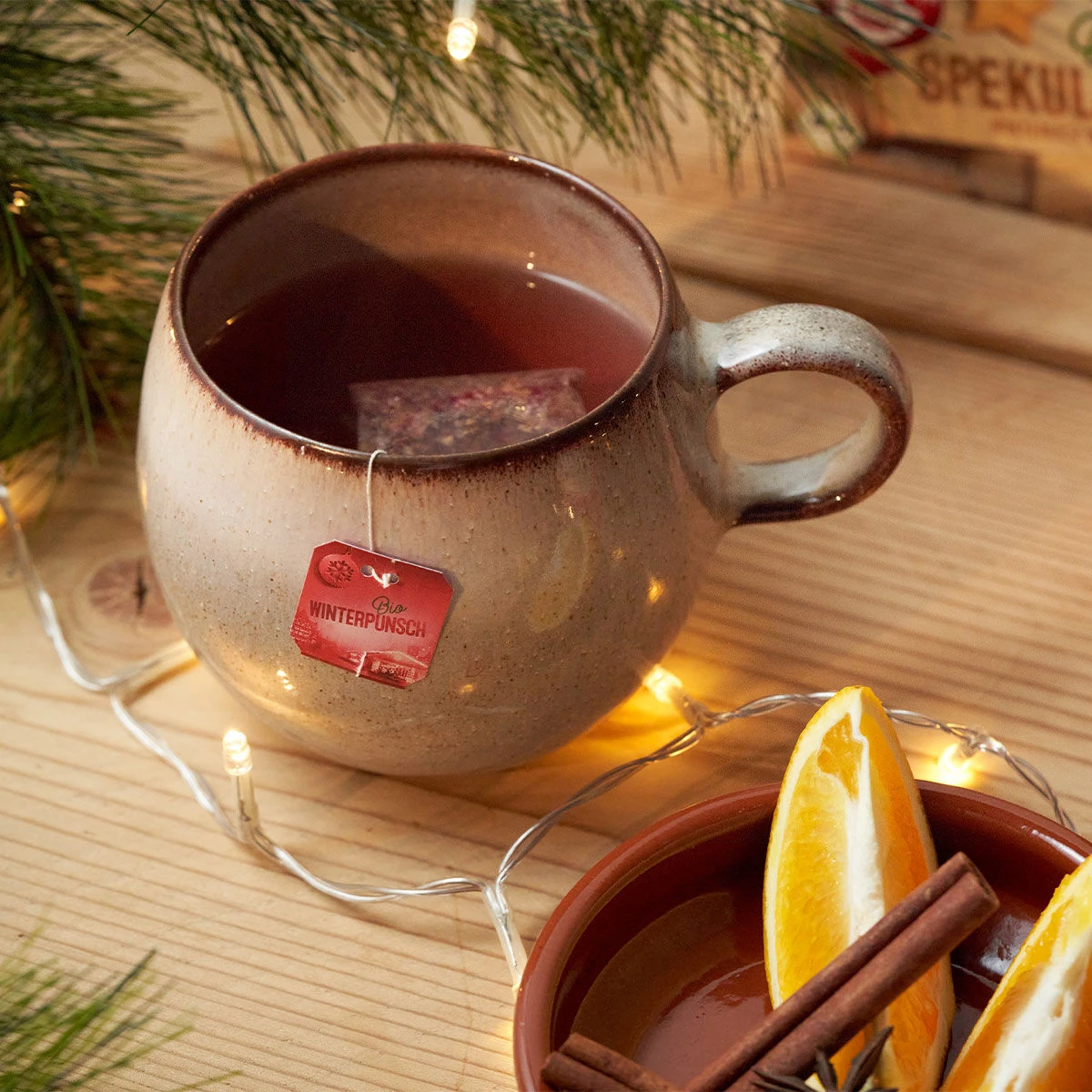 Eine Keramiktasse Bio-Winterpunsch-Tee mit einem „Winterpunsch“-Teebeutel steht auf einem Holztisch, umgeben von Tannennadeln, Lichterketten, getrockneten Orangenscheiben und Zimtstangen.