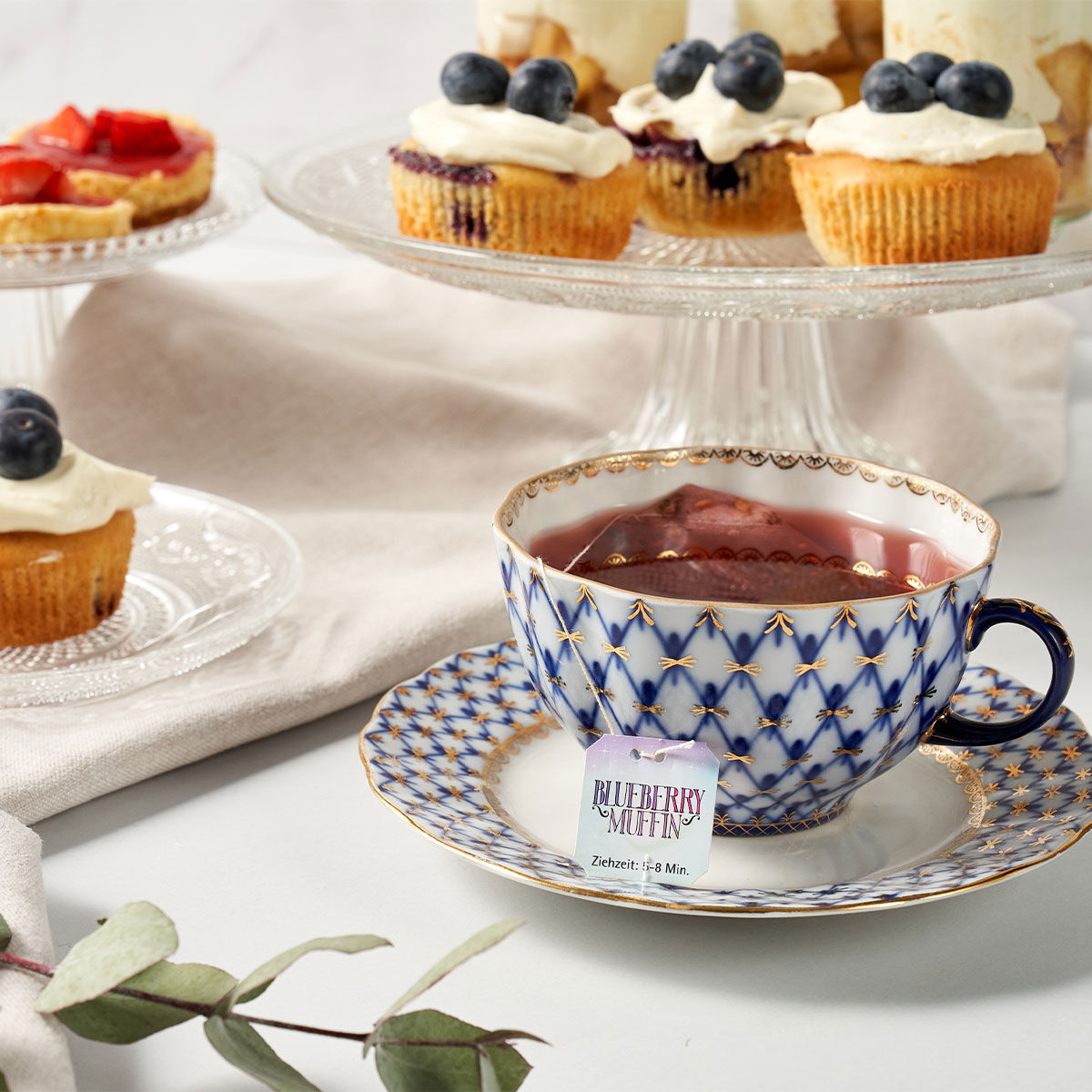 Eine gemusterte Teetasse mit Tee steht auf einer passenden Untertasse neben einem Sweeteas „Blueberry Muffin“-Teebeutel. Im Hintergrund sind Cupcakes mit Zuckerguss und Blaubeeren auf Glasständern ausgestellt.